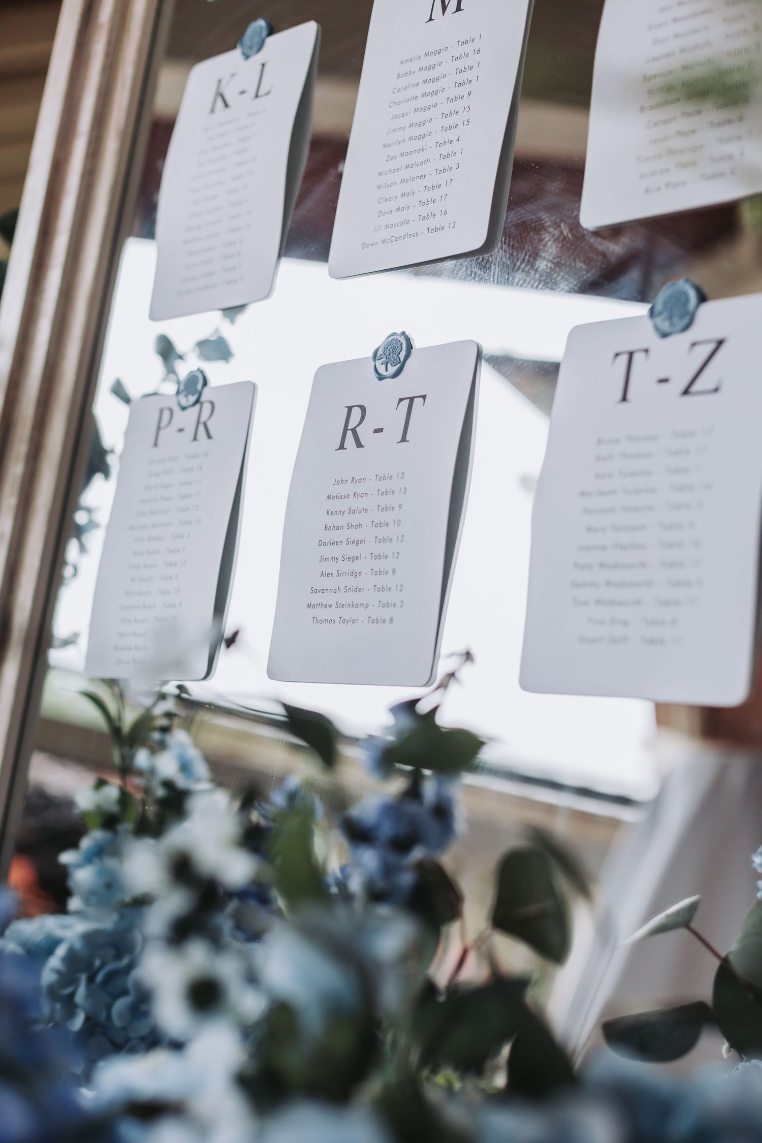 Elegant wedding seating chart cards arranged alphabetically on mirror display with blue floral arrangement below.