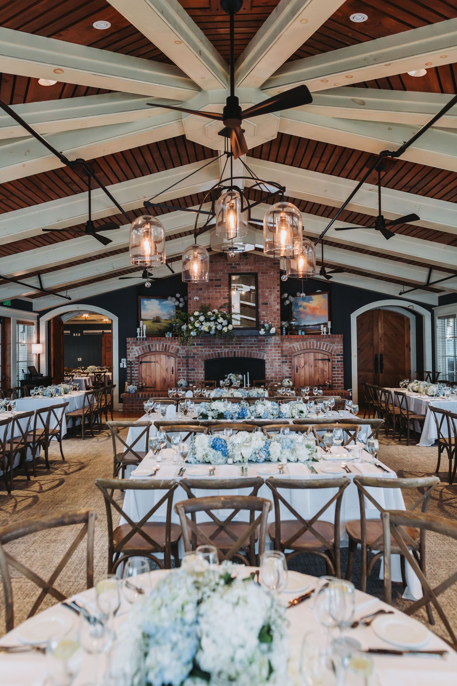 A stunning wedding reception venue featuring vaulted wood ceilings, a brick fireplace, and blue hydrangea centerpieces.