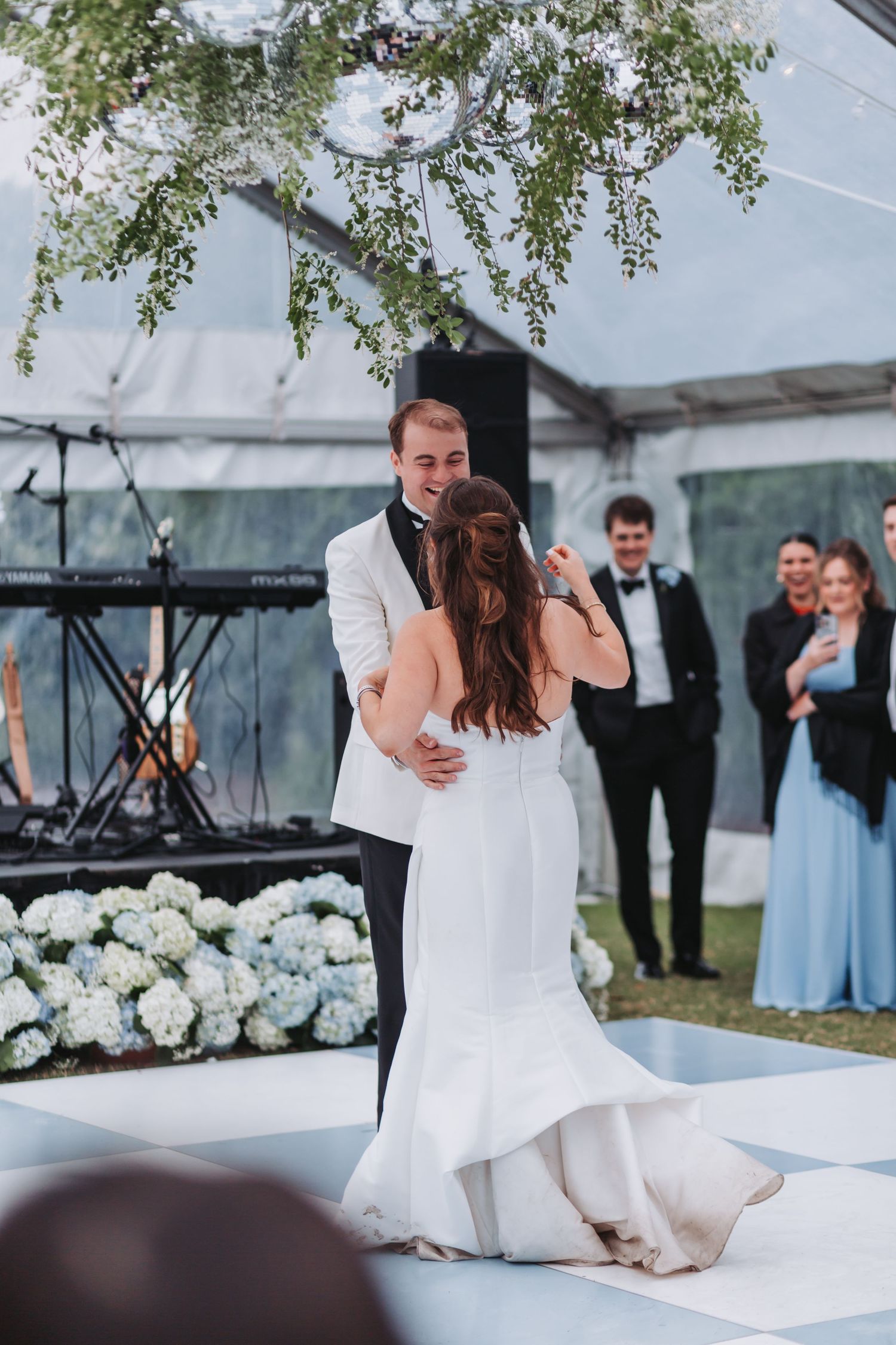 Groom in white tuxedo sharing first dance with bride on white dance floor under tent.
