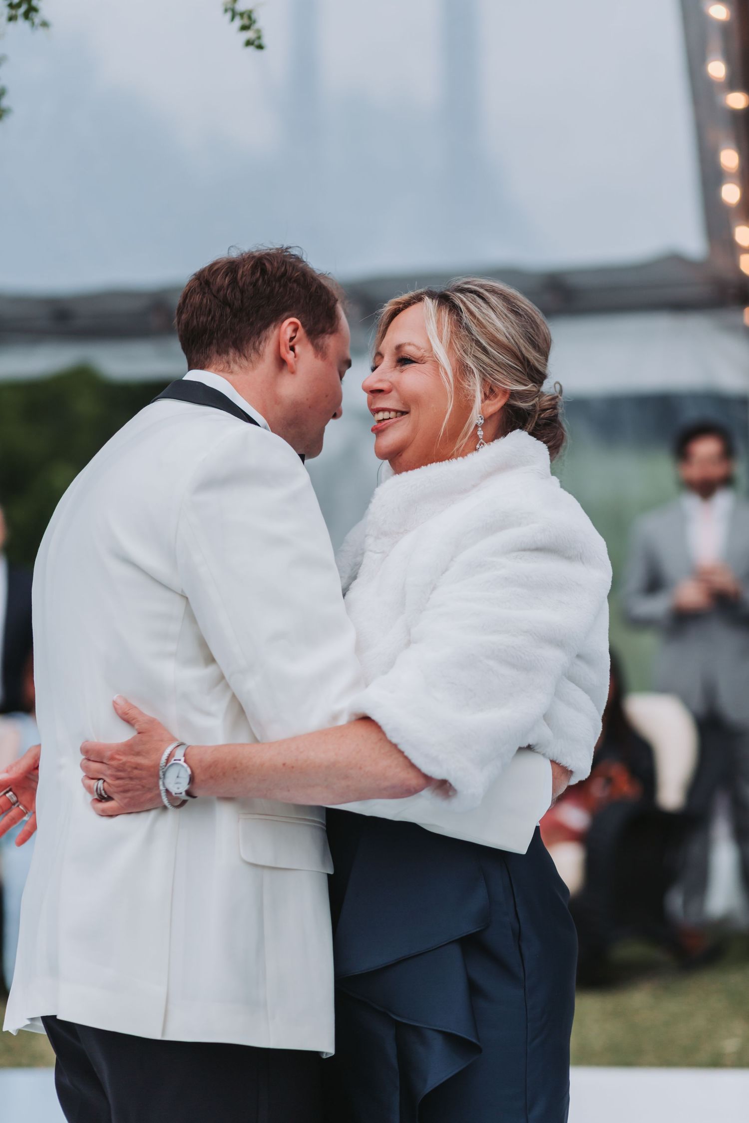 Groom in white tuxedo shares tender dance with smiling woman in white fur wrap at wedding reception.