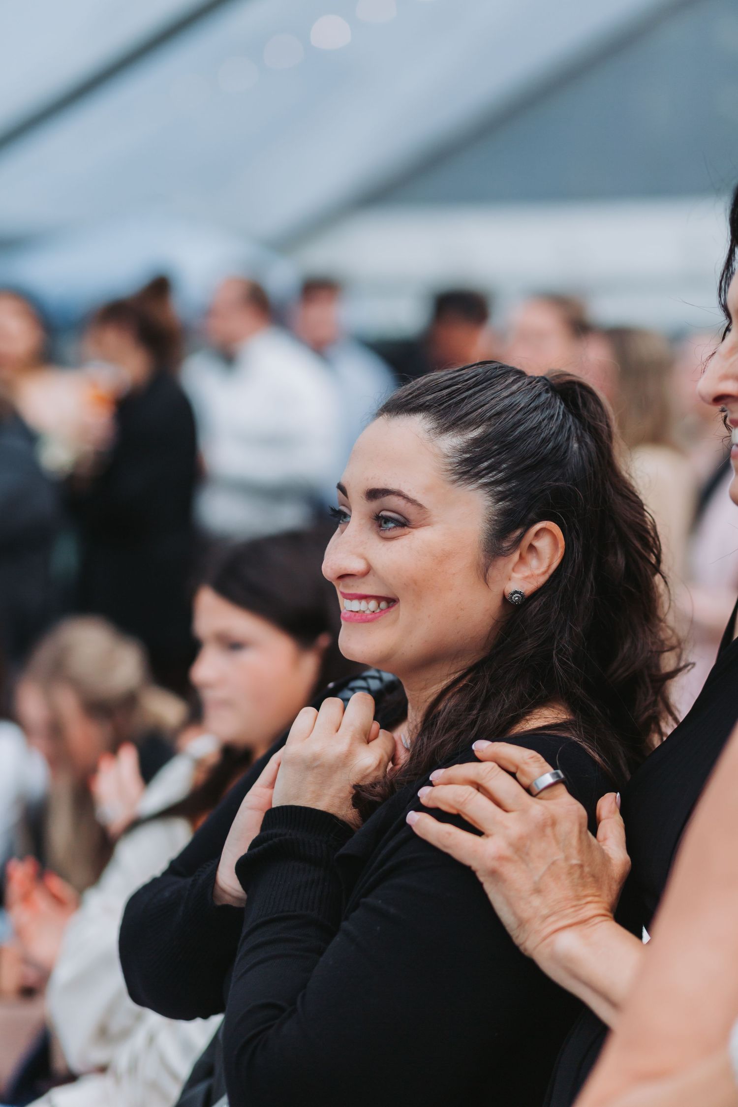 Smiling dark-haired woman in black dress watches emotional moment at wedding ceremony in white tent.