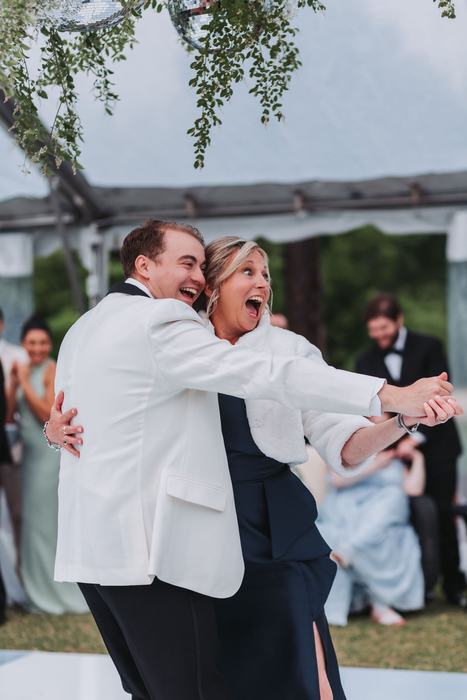 Groom in white jacket joyfully lifts a woman in a navy dress during an outdoor wedding celebration.