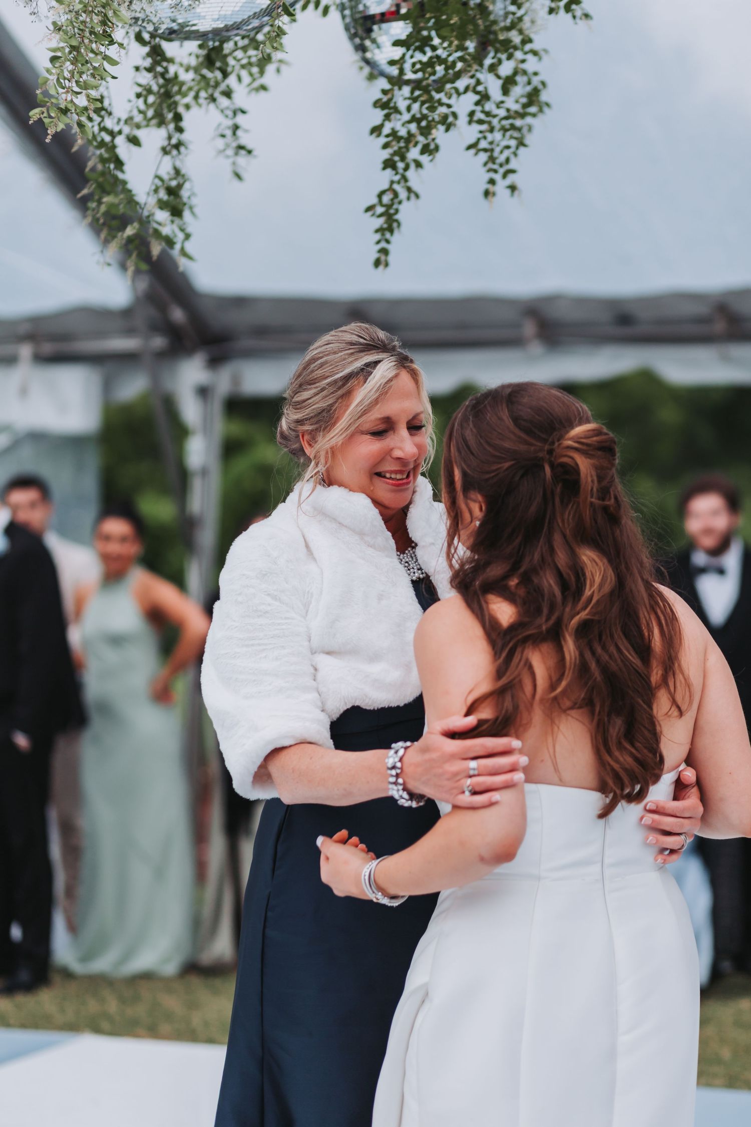 Mother embracing bride in strapless gown on dance floor under greenery at tented wedding reception.