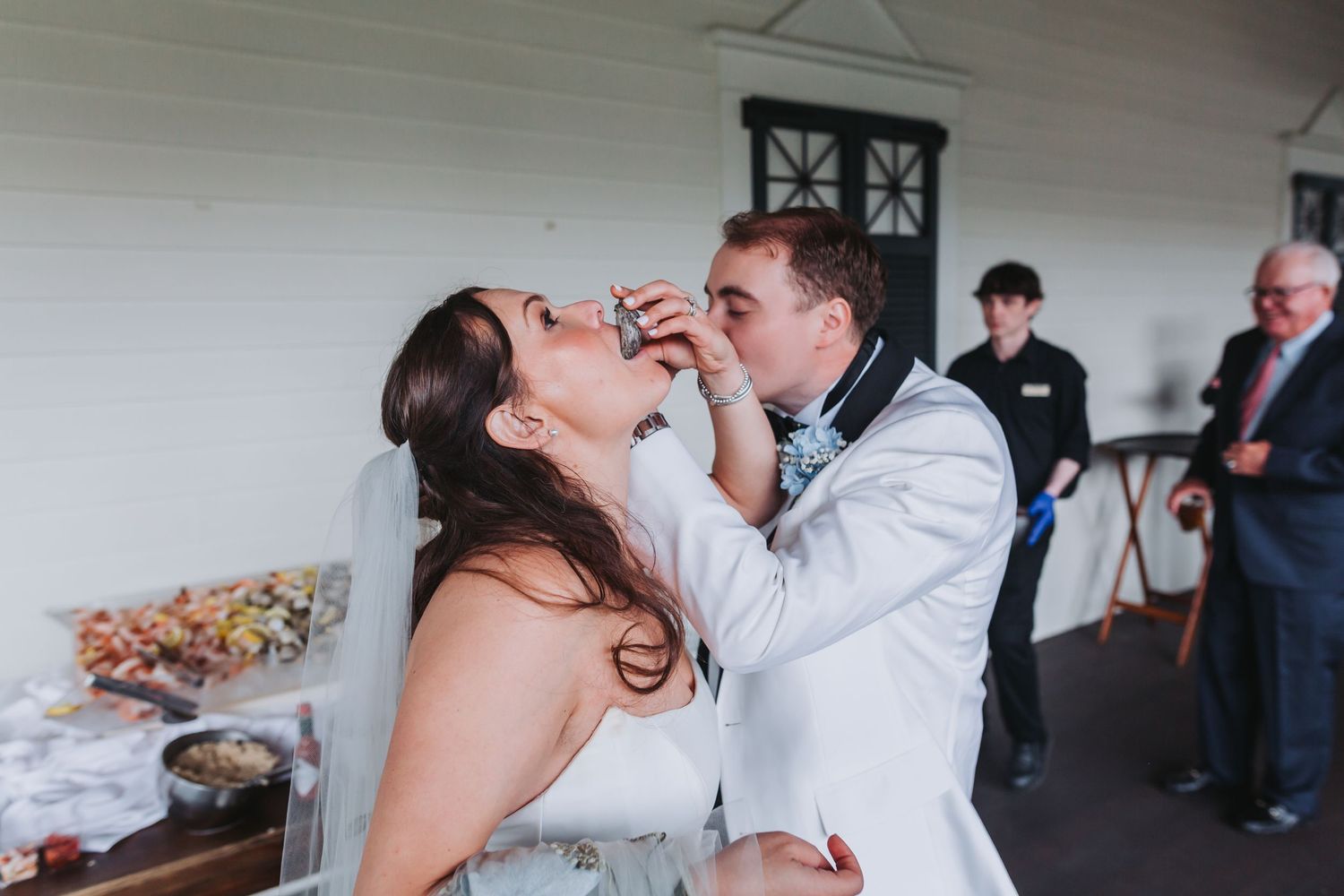 Bride and groom playfully feed each other wedding cake on venue porch during reception celebration.