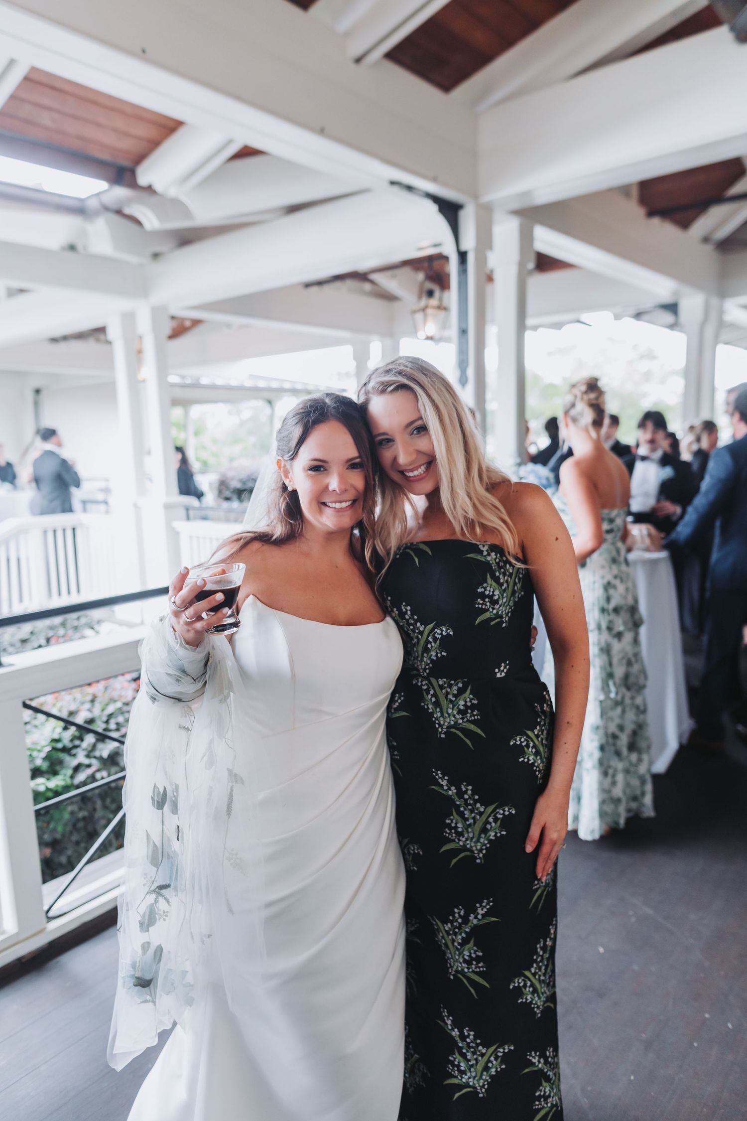 Bride in white gown posing with guest in black floral dress at outdoor wedding cocktail hour.