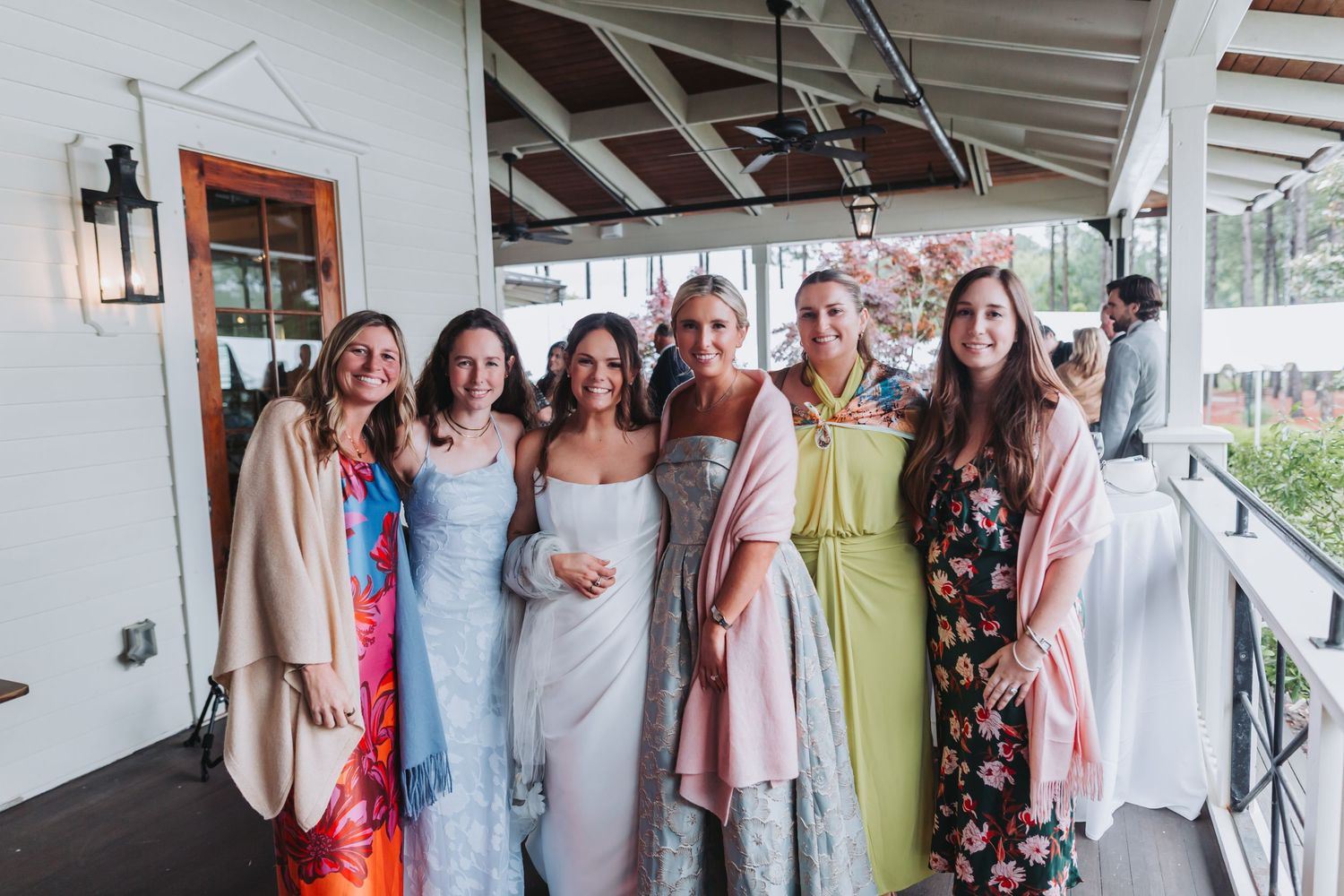 Bride poses with six female guests in colorful dresses and wraps on a covered outdoor porch at wedding.