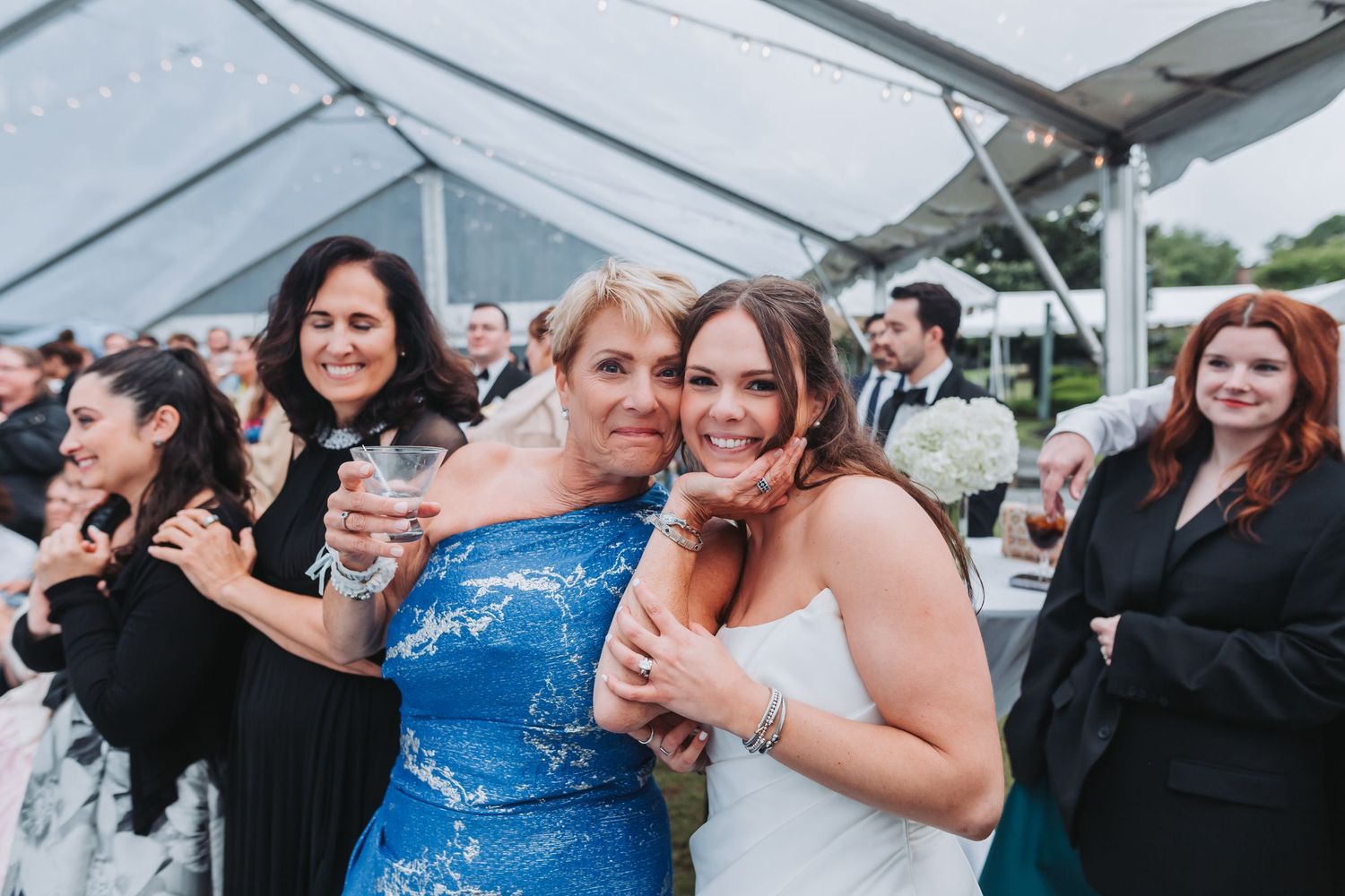 A bride in a white gown hugs a woman in a blue sequin dress at a wedding reception under a tent.