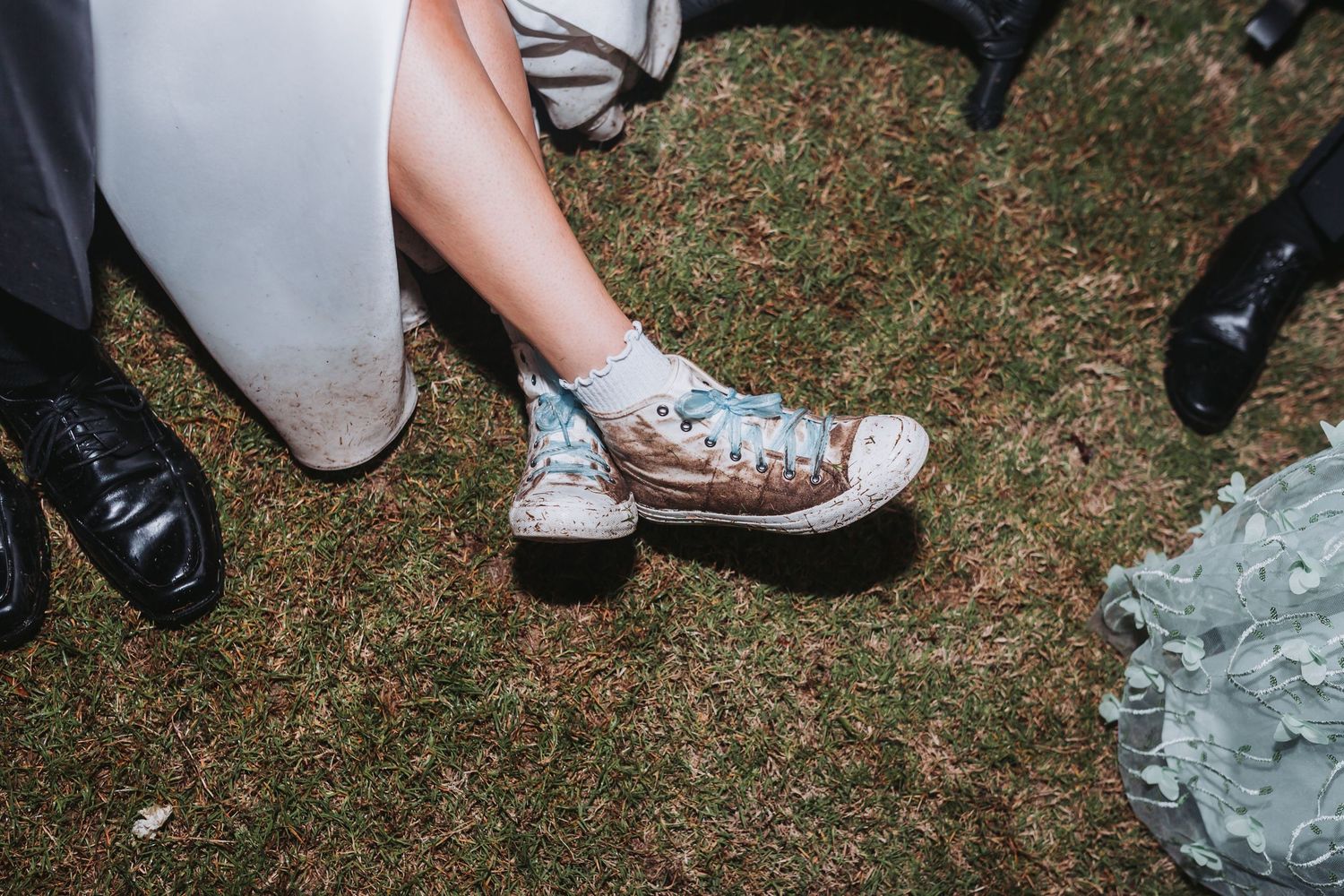A bride wearing worn blue Converse sneakers sits on grass surrounded by wedding guests in formal shoes.