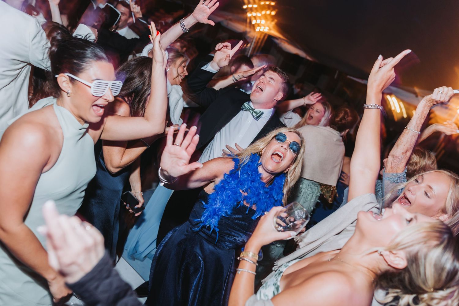 Guests dancing energetically at wedding reception with woman in blue feather boa celebrating on dance floor.