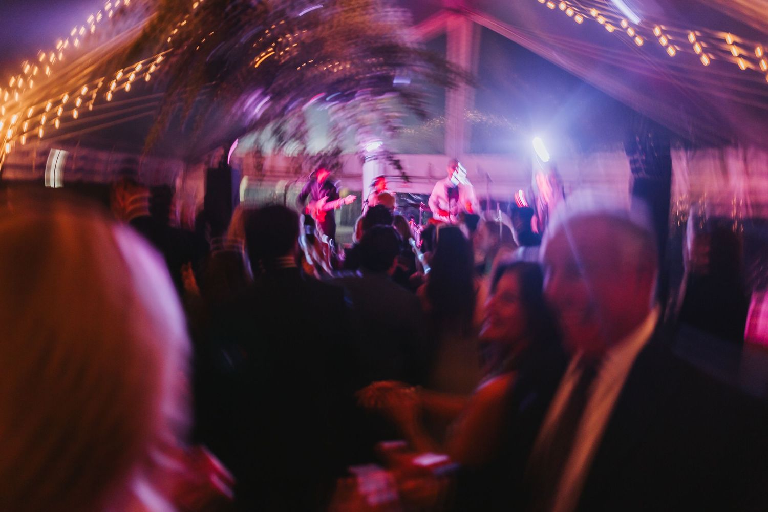 Blurred motion shot of wedding reception guests dancing under warm string lights in a tent venue.