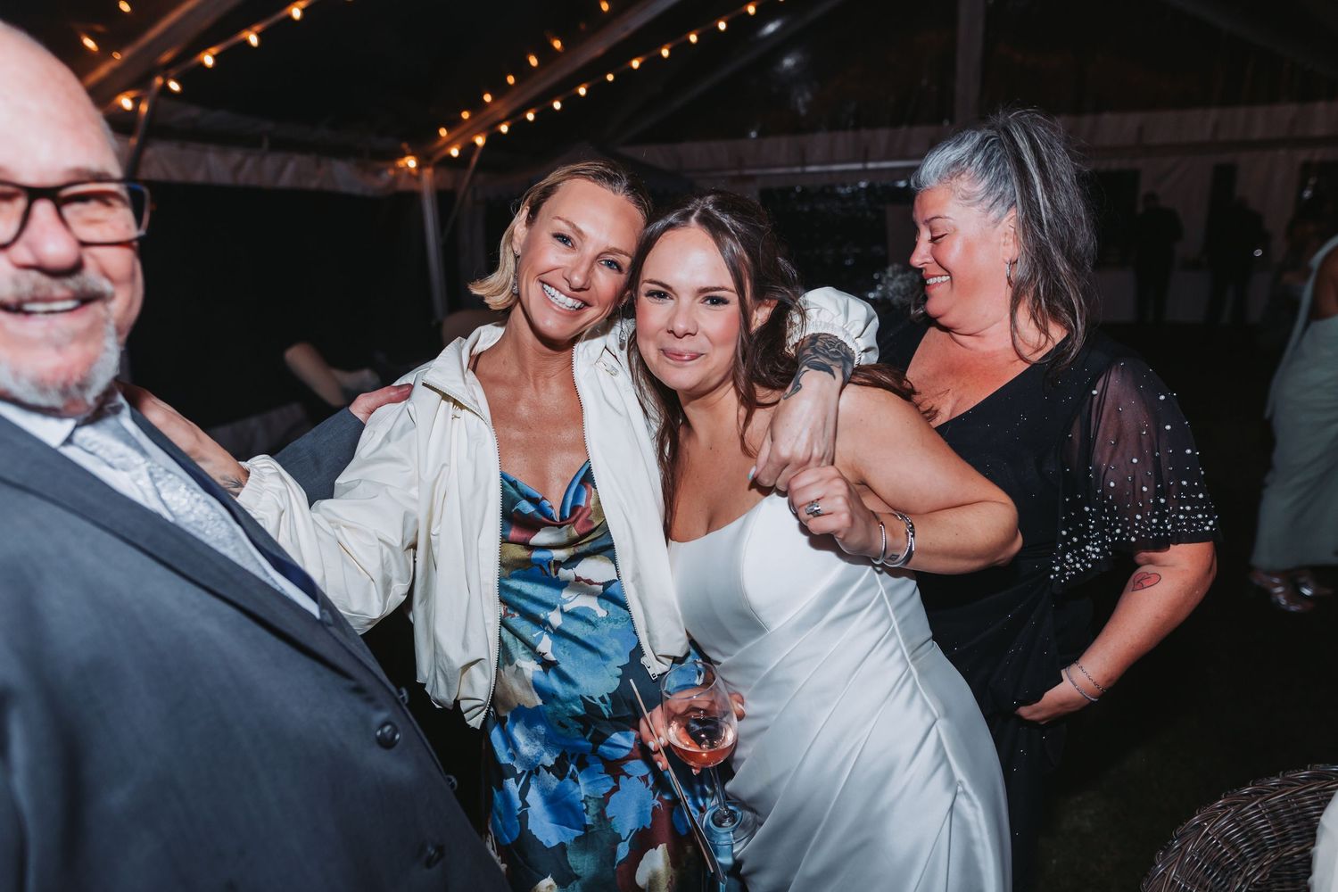Bride in white dress laughs and dances with guests at lively outdoor wedding reception under string lights.