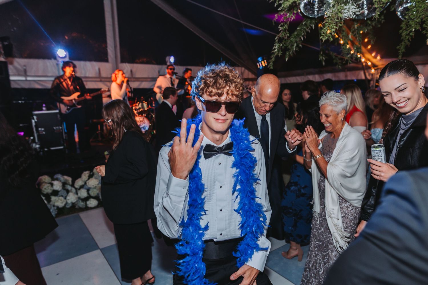 Young man in tuxedo with blue feather boa and sunglasses dances at elegant wedding reception party.