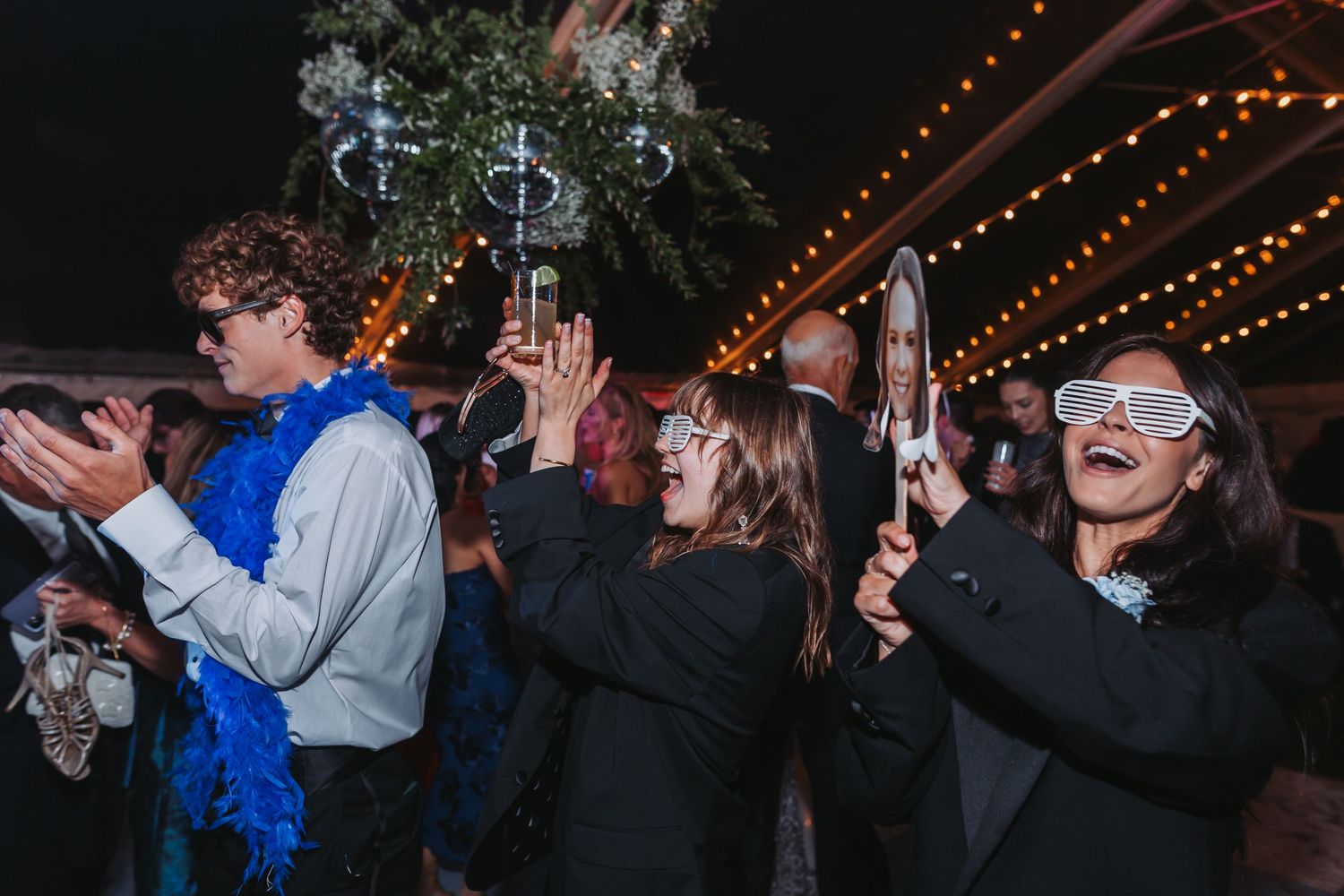 Wedding guests dance energetically at night under string lights wearing fun accessories.