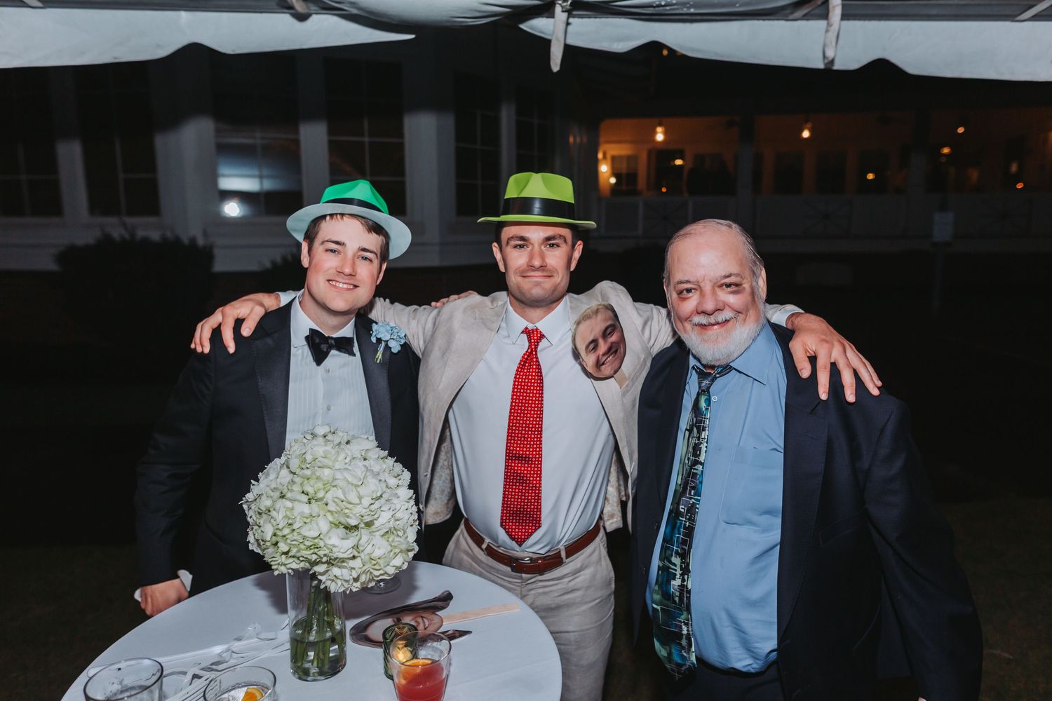 Three smiling men pose together at an outdoor wedding reception, two wearing festive green hats.