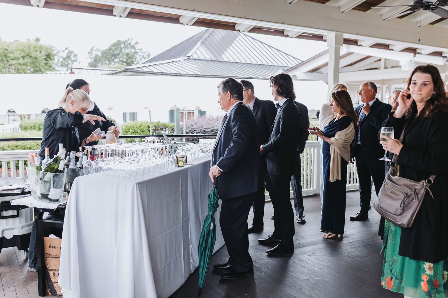 Guests mingle at an outdoor wedding cocktail hour bar with champagne glasses and wine bottles on a covered patio.