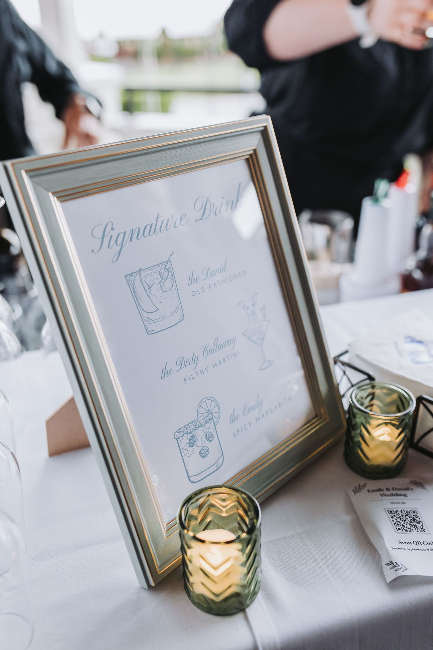 Framed wedding signature drinks menu displayed on a table with green glass candle holders at an elegant reception.