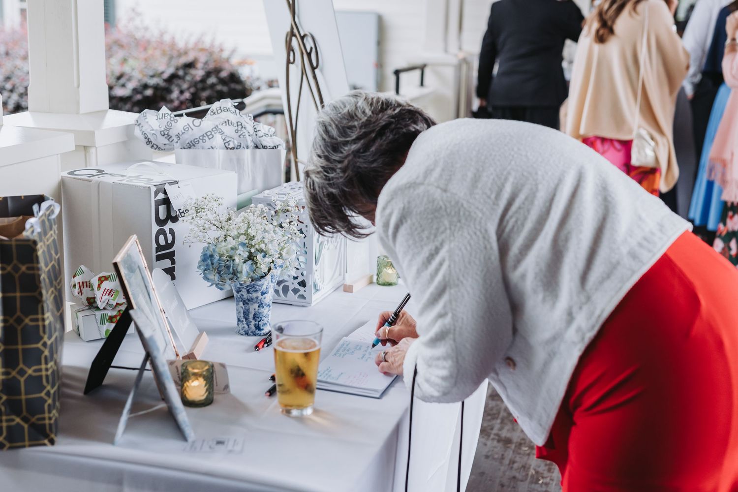 A guest in red pants signs the wedding guest book at a decorated reception table.