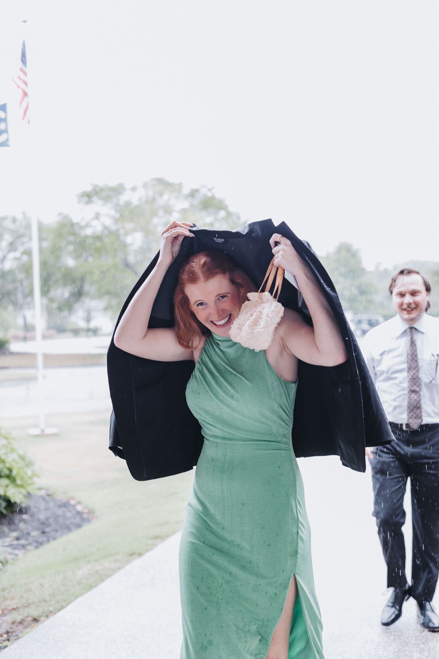 Smiling wedding guest in green dress uses black suit jacket as umbrella shield in the rain.