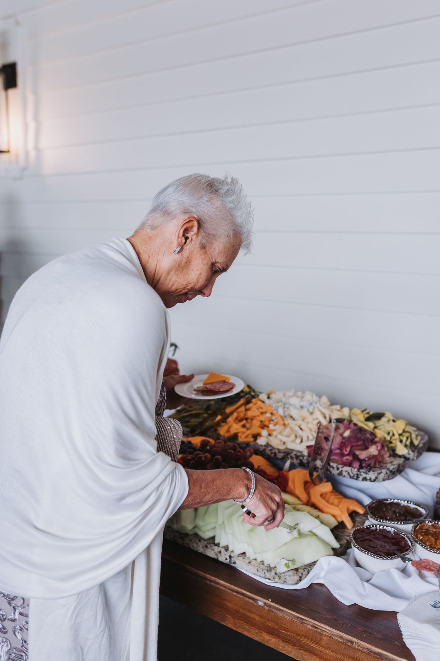 Elderly woman with white hair selects food from elaborate grazing table spread with fruits and cheeses.