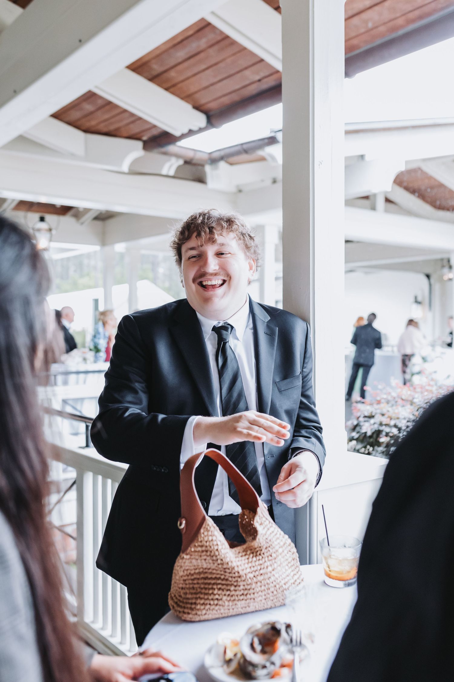Laughing man in black suit and tie at wedding reception standing near wicker handbag on white table.