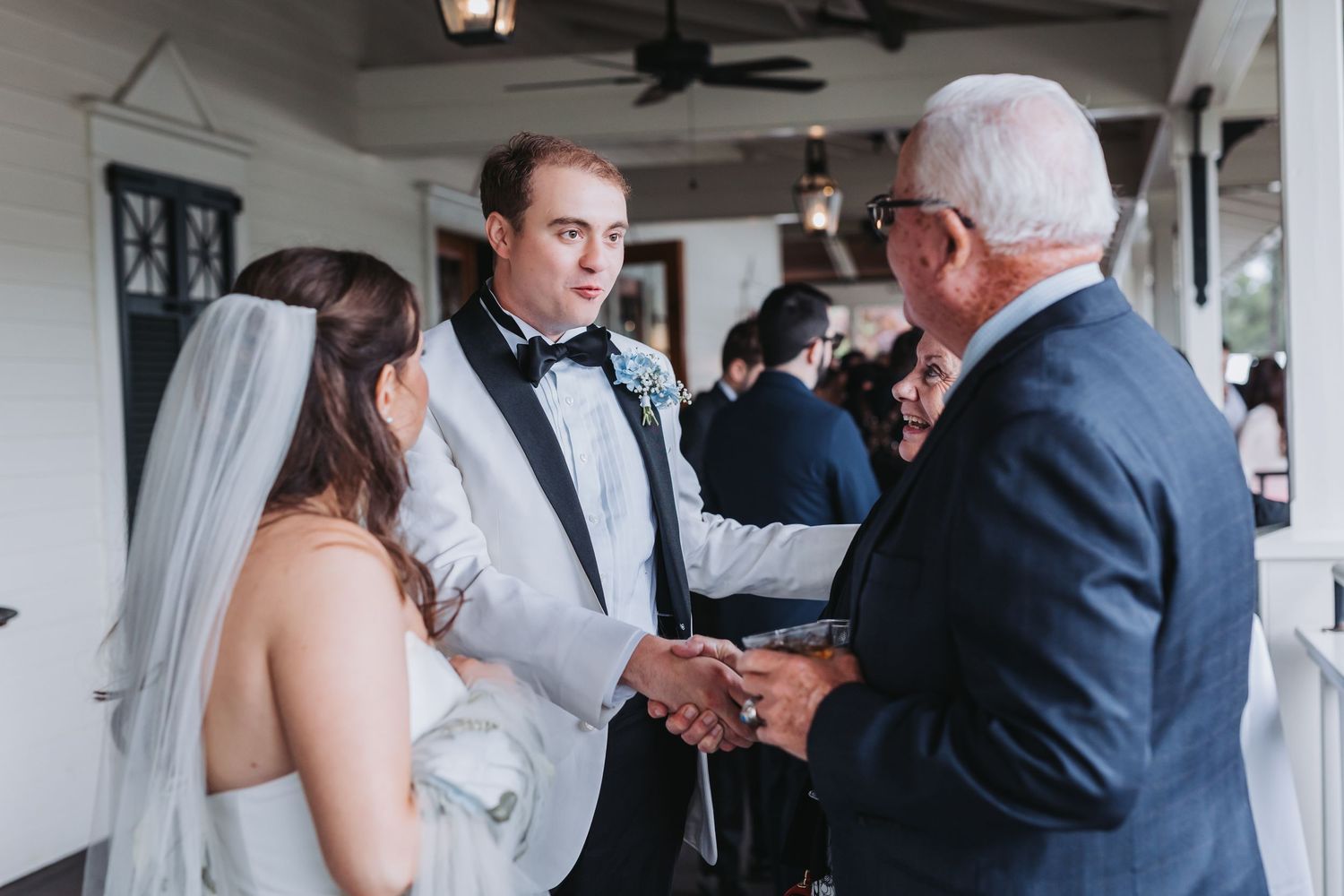 Bride and groom greet an elderly guest with a handshake on a porch after their wedding ceremony.
