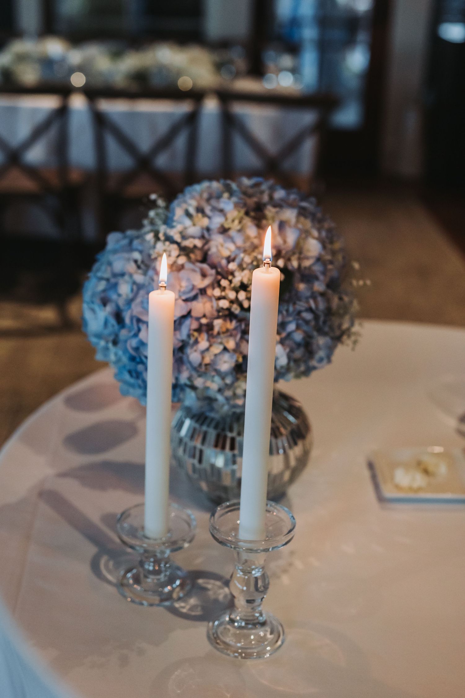 Two lit white taper candles in crystal holders beside a blue hydrangea floral centerpiece on a wedding table.