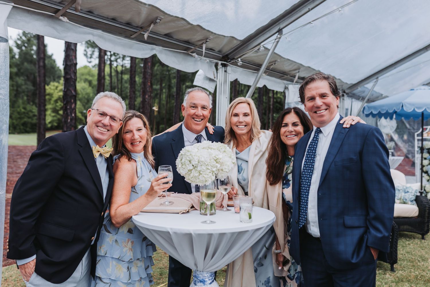 Group of six smiling wedding guests posing together at an outdoor tented reception cocktail hour.