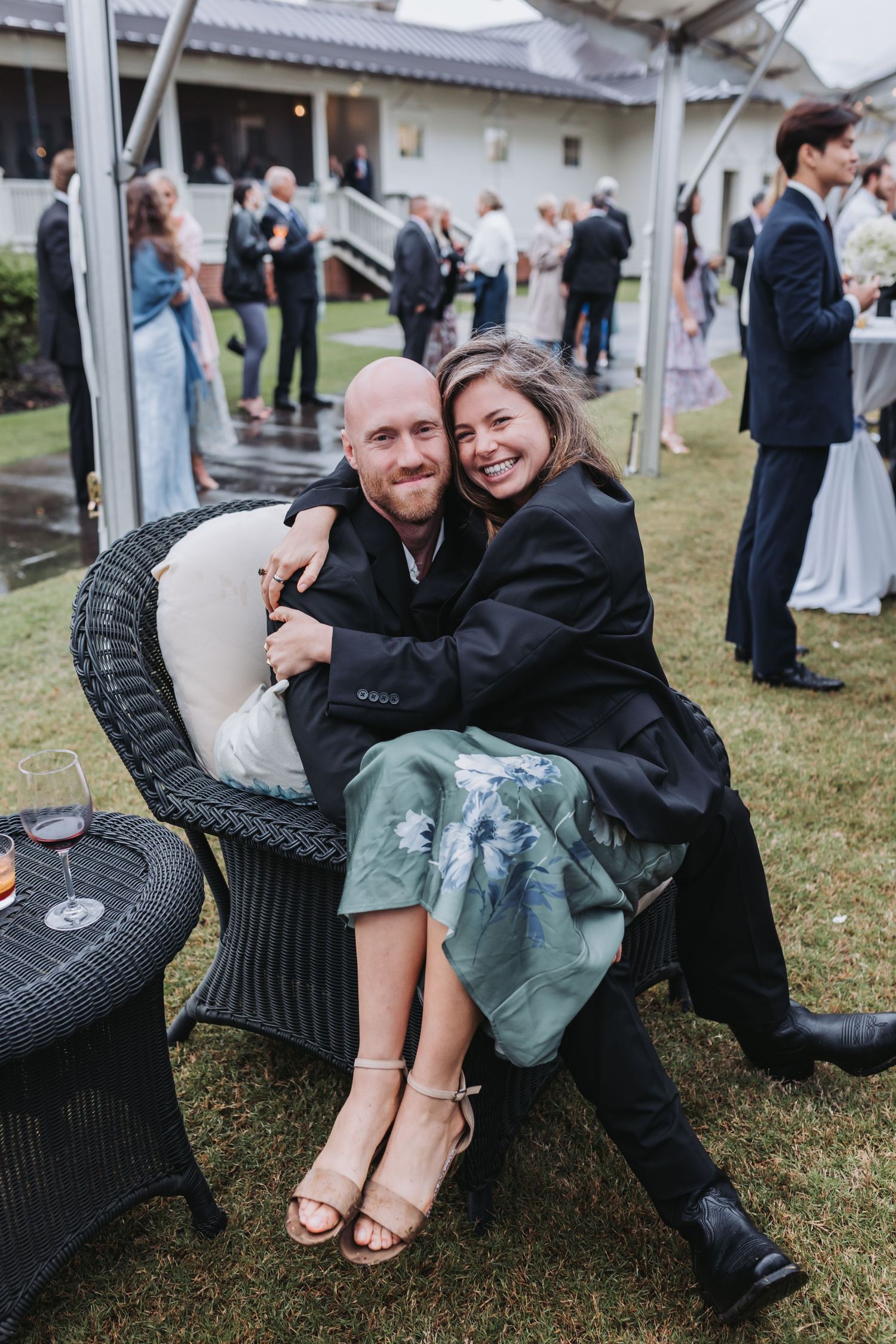 A smiling couple cuddles together in a wicker chair at an outdoor wedding reception on a lawn.