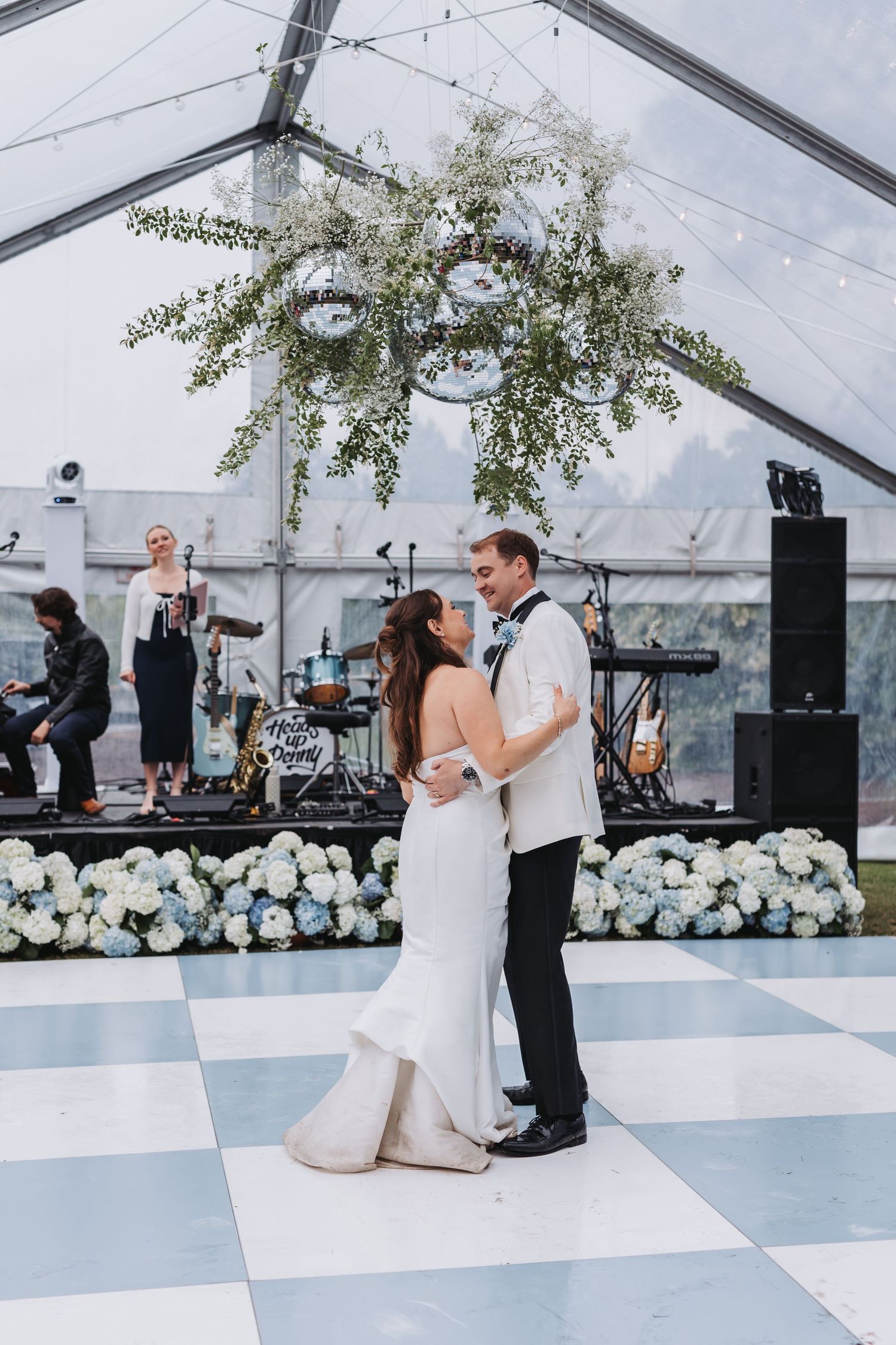 Bride and groom share first dance on checkered floor under floral chandelier with live band performing behind them.