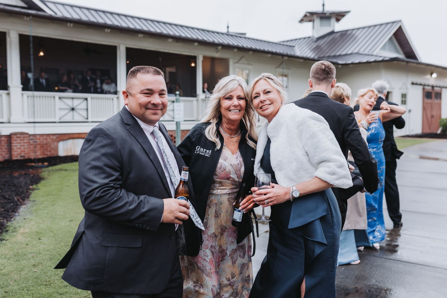 Three wedding guests smile and pose together outside a white barn venue during a rainy outdoor wedding reception.