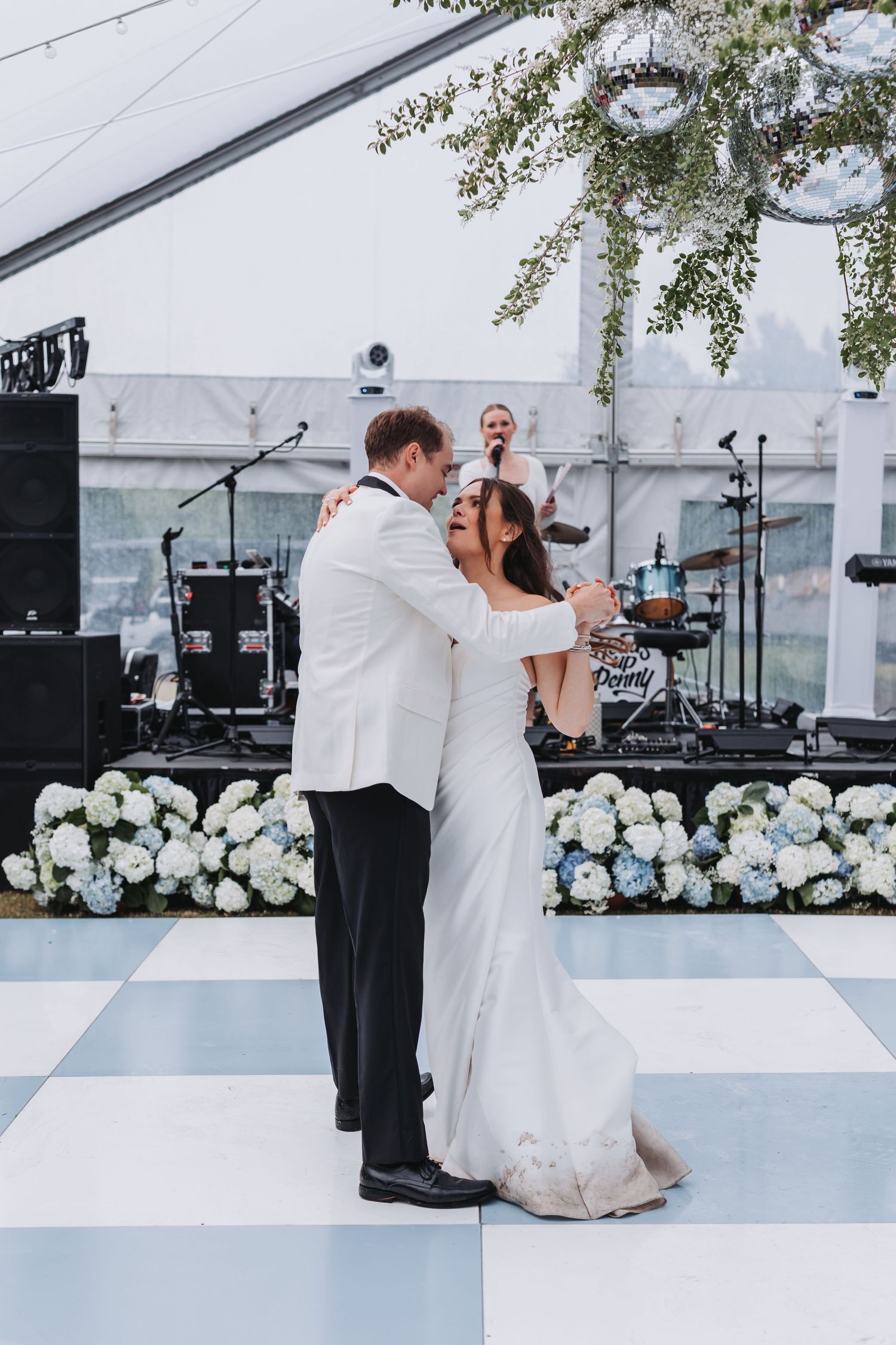 Newlyweds share an intimate first dance on blue and white checkered floor with hydrangea floral wall backdrop.