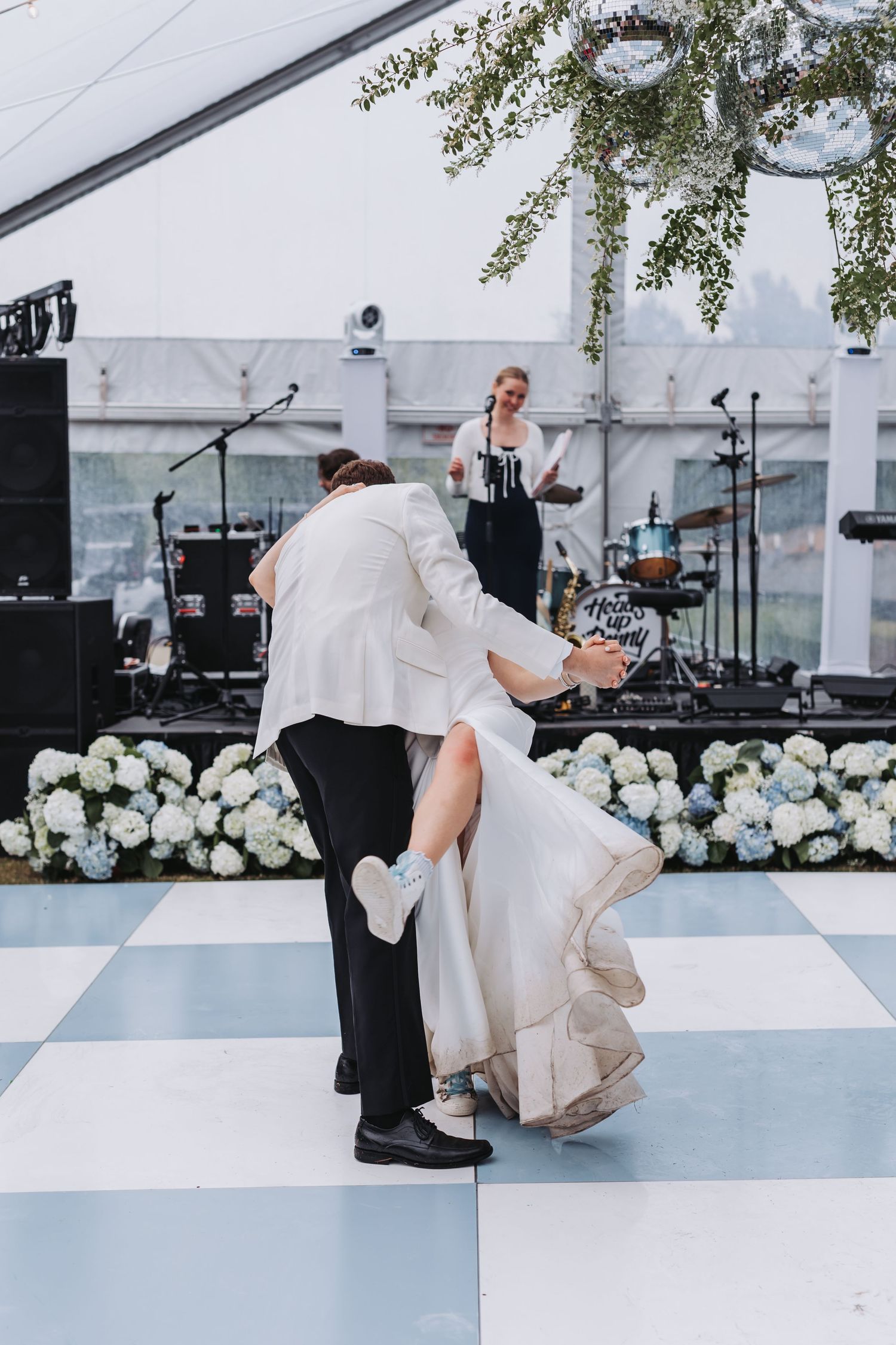 Groom in white jacket dips bride in wedding gown on checkered dance floor during first dance.