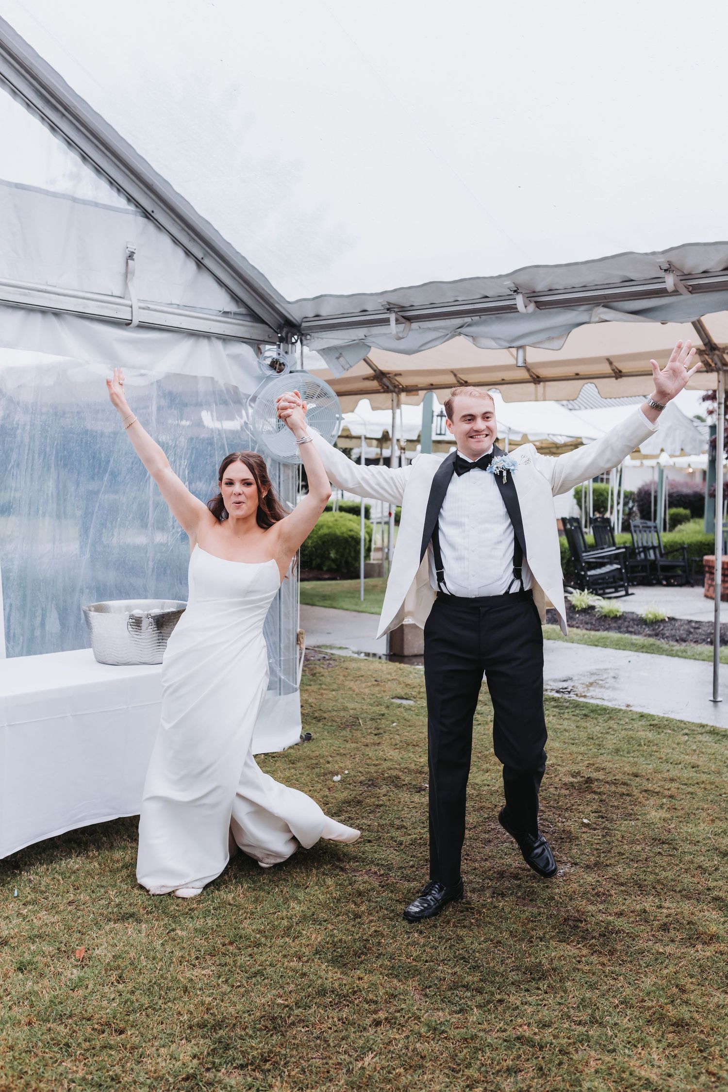 Joyful bride and groom raising arms in celebration under a clear tent at their outdoor wedding reception.