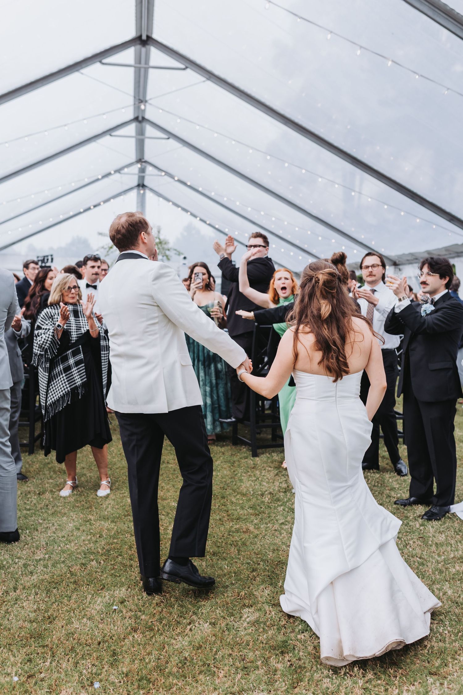 Bride and groom enter reception holding hands as cheering guests celebrate under a clear tent on a grassy lawn.