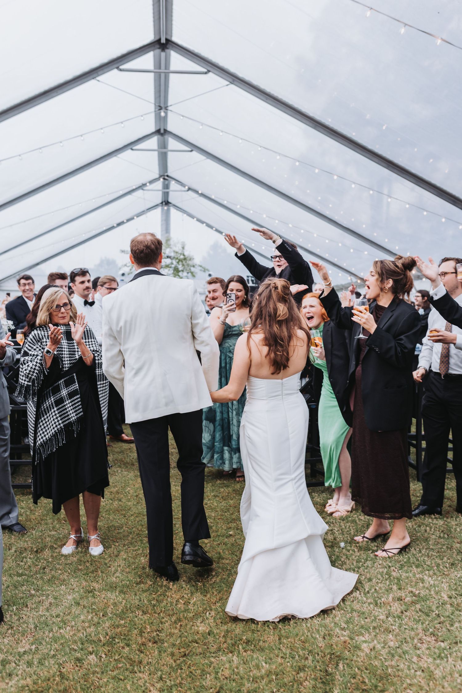 Bride and groom entering their wedding reception under a clear tent surrounded by cheering guests.