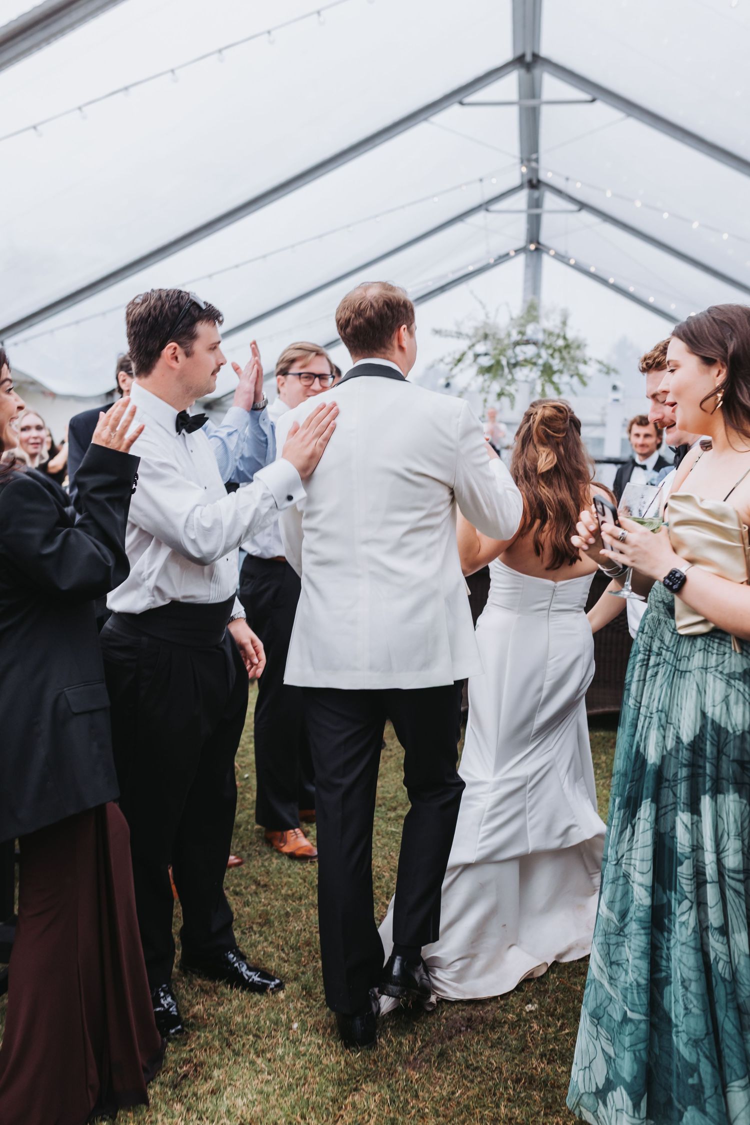 Wedding guests cheer as bride and groom in white jacket enter reception under a clear tent with string lights.