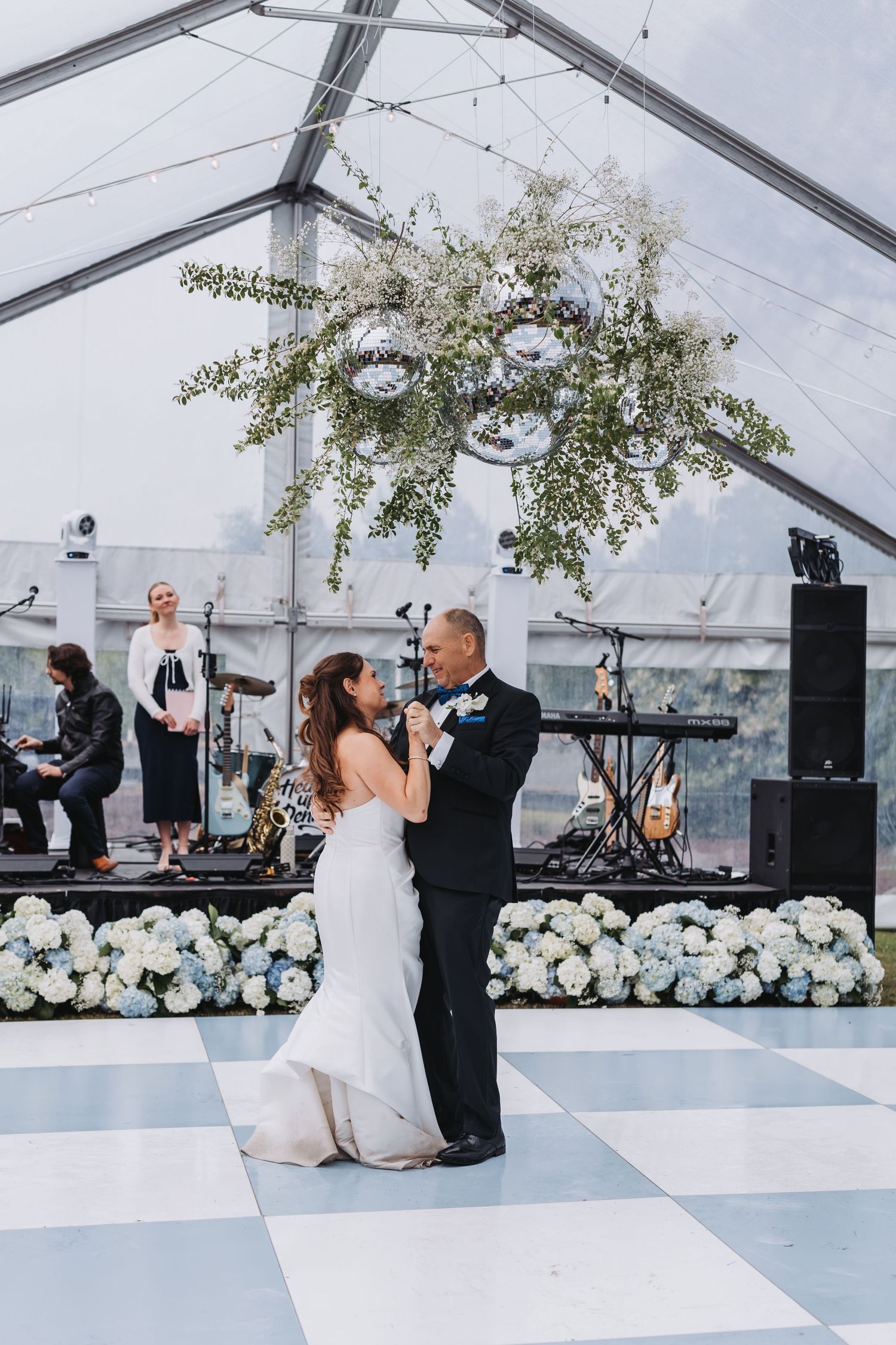 Couple shares first dance under a floral chandelier on a checkered floor in a clear tent.