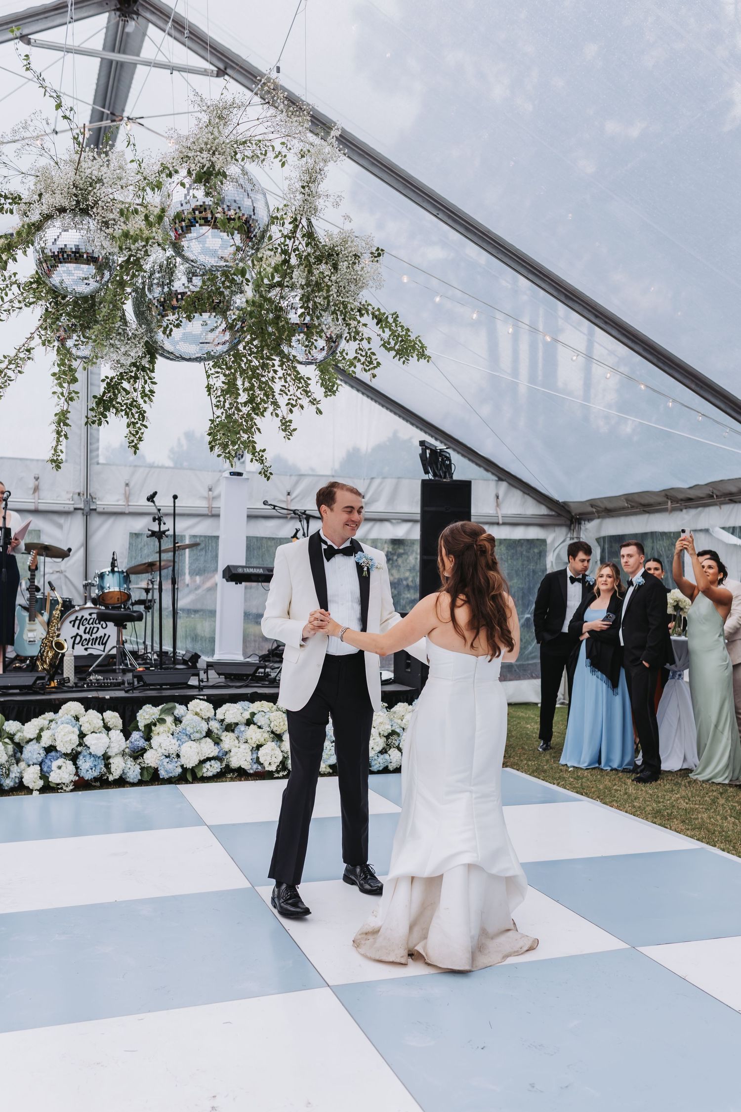 Bride and groom share first dance on checkered floor as wedding party watches under floral chandelier.