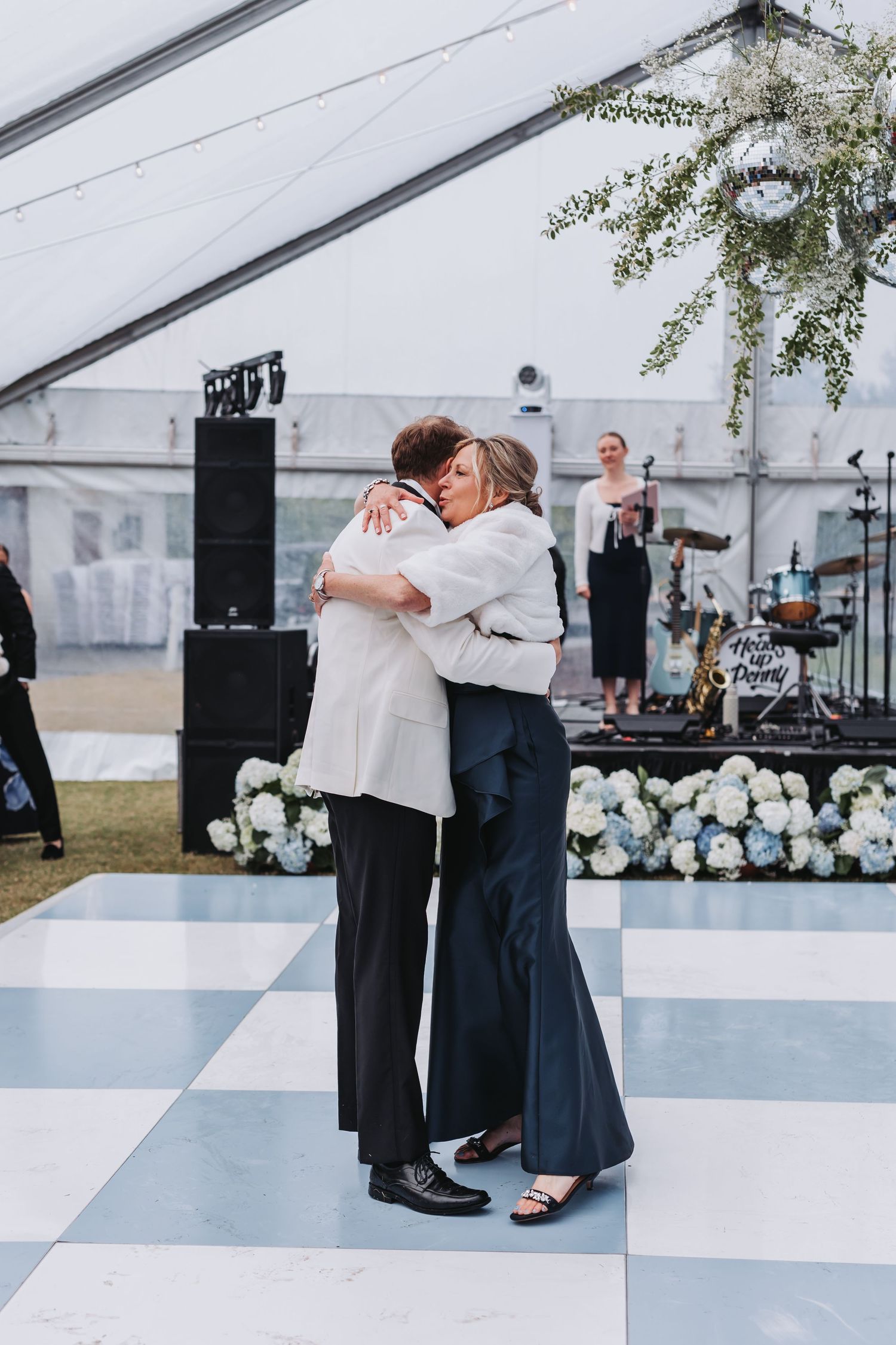 Couple sharing emotional hug on checkered dance floor under floral chandelier at elegant wedding reception.