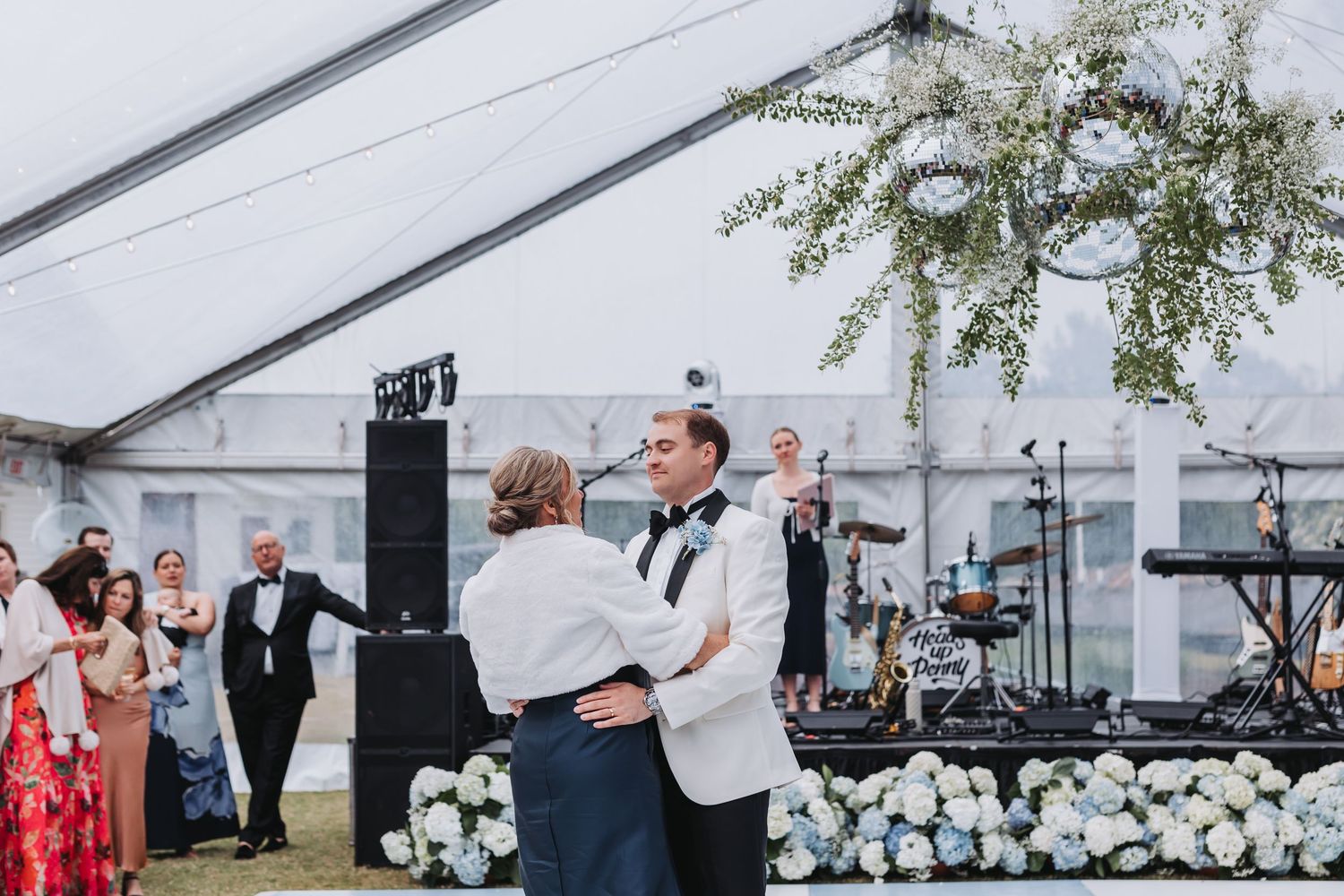 Groom dances with mother on checkered dance floor surrounded by white hydrangea arrangements at wedding reception.