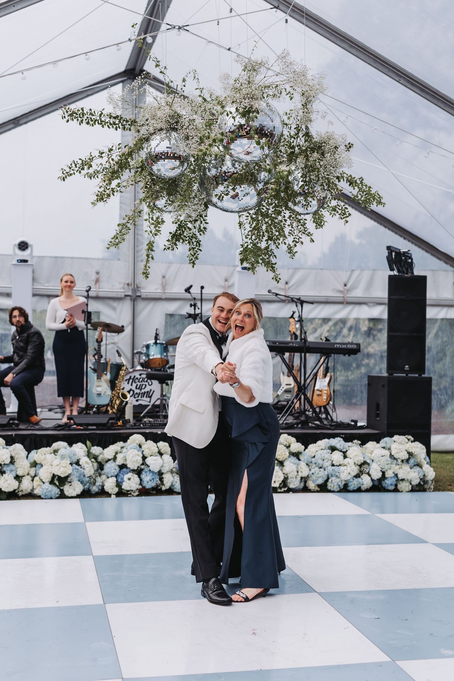 Couple dancing joyfully on checkered floor under hanging greenery with live band performing at wedding.