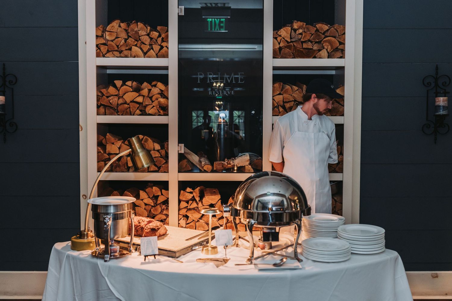 Catering staff stands behind an elegant buffet table with silver chafing dishes and stacked white plates.