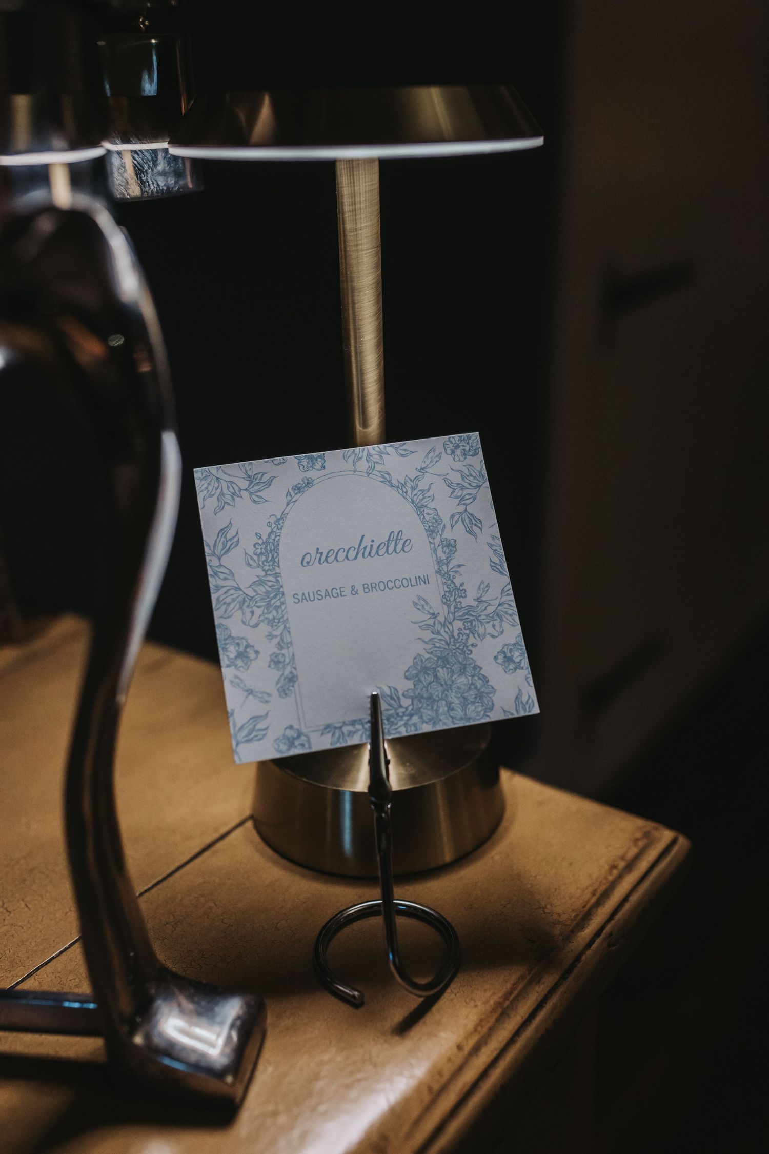 Elegant wedding menu card for orecchiette with sausage and broccoli displayed in a metal holder beside a lamp.