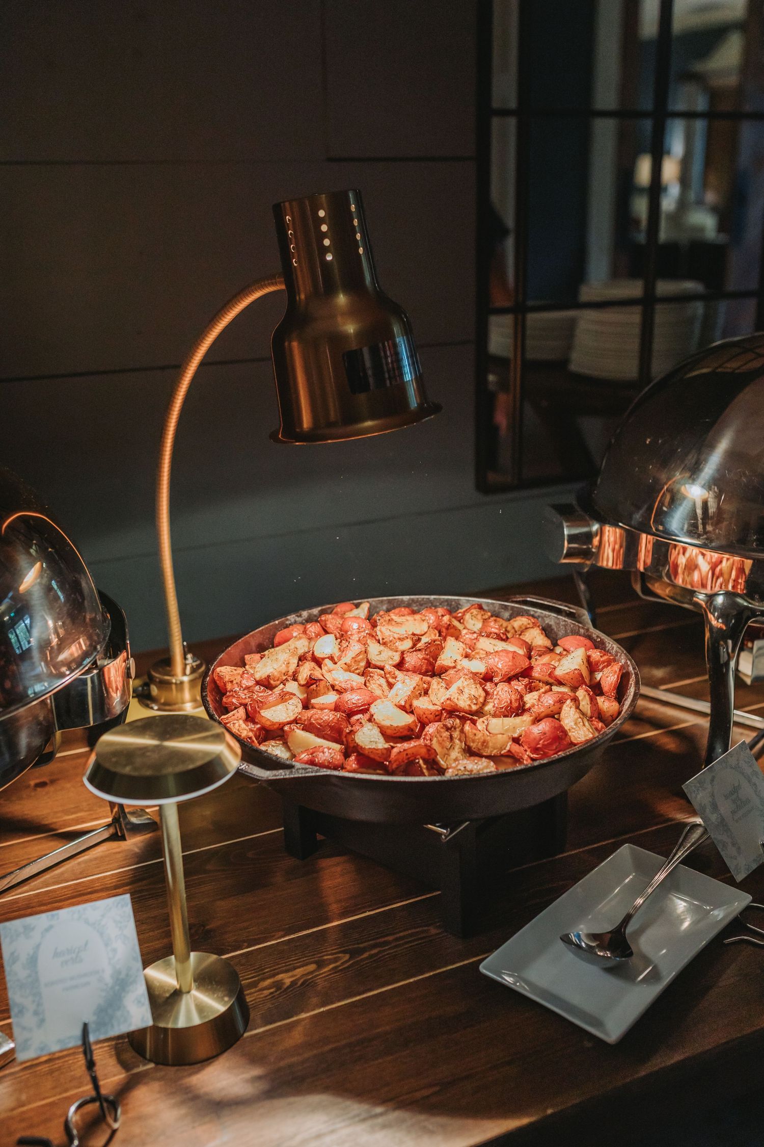 A warm buffet station with roasted potatoes in a pan under a brass heat lamp.