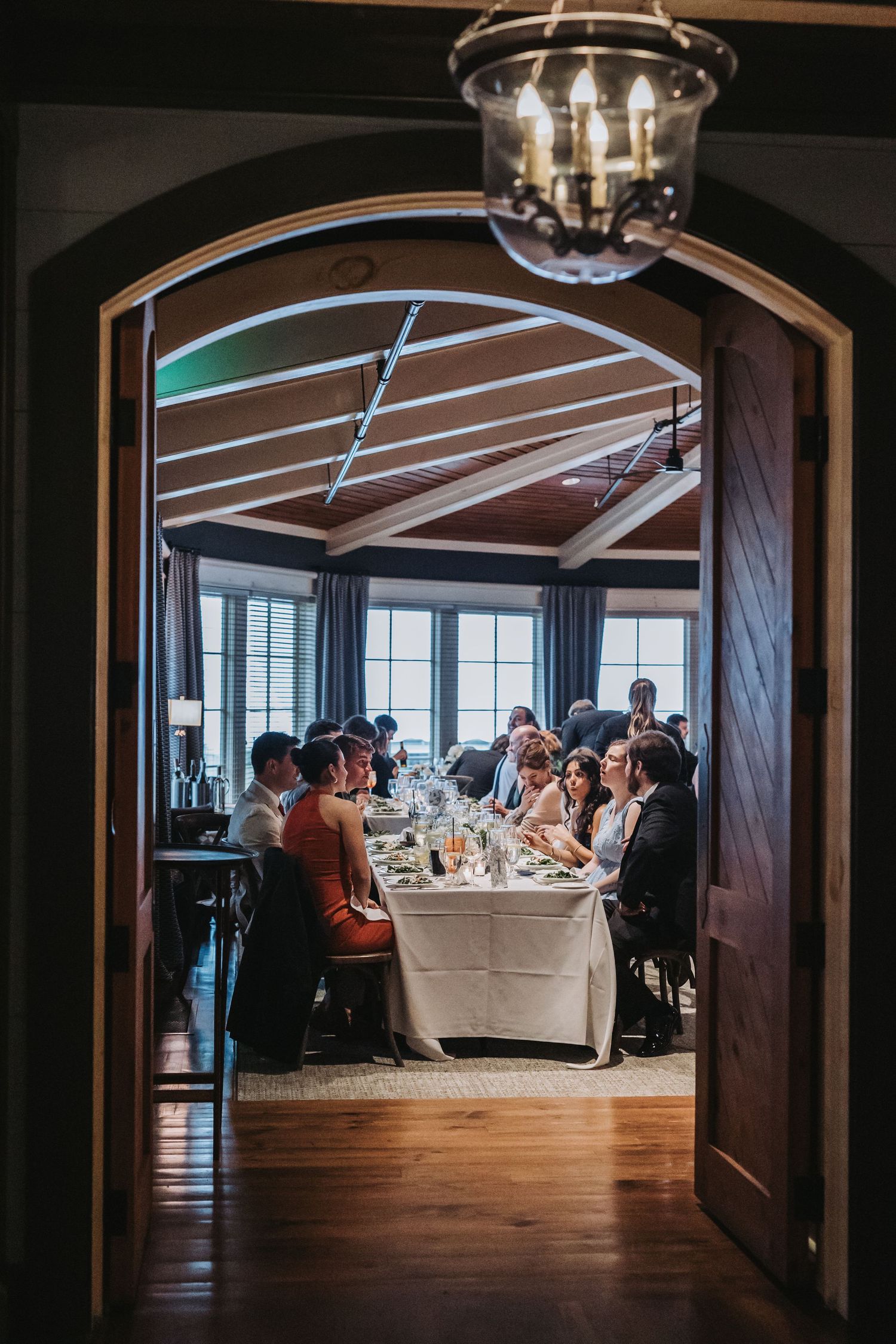 Romantic wedding dinner reception viewed through arched wooden doors with guests seated at long table.