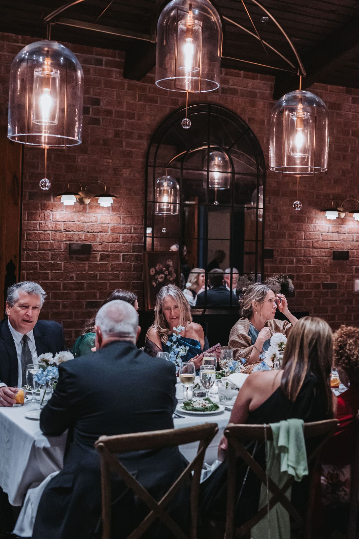 Wedding guests dining at round tables under warm pendant lights in a brick-walled reception venue.