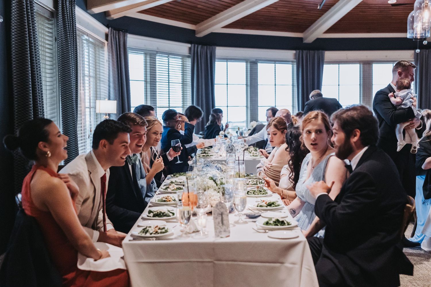 Wedding guests seated at elegant long dining table enjoying dinner conversation in a bright windowed venue.