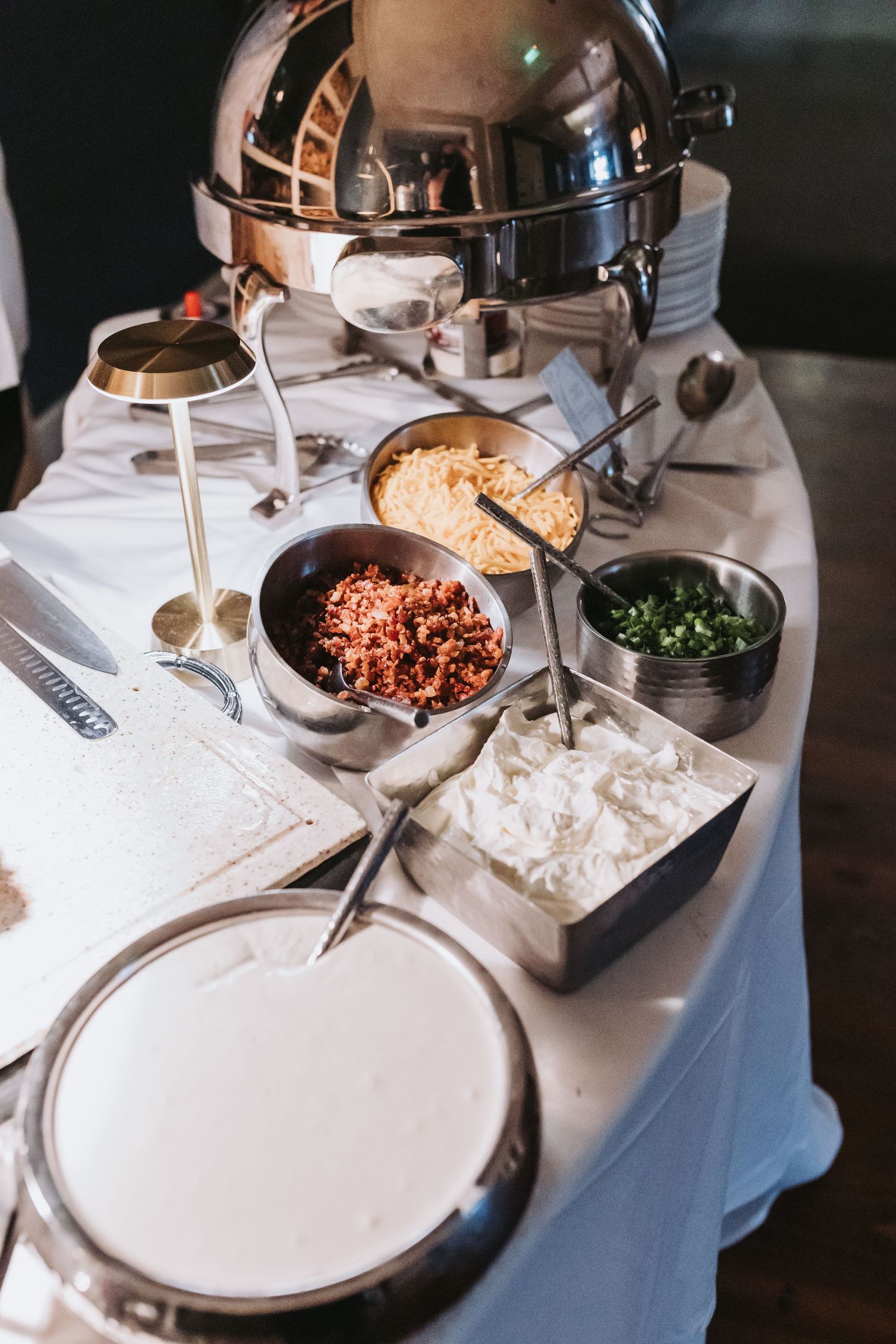 Elegant wedding buffet table with chafing dishes, toppings, and condiments on white linen.