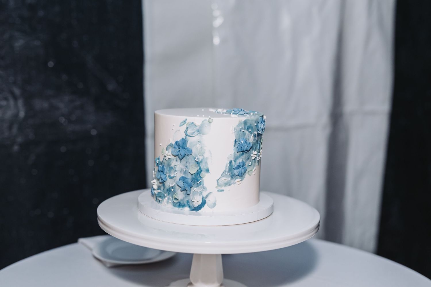 Elegant single-tier white wedding cake decorated with blue watercolor floral brushstroke design on a white stand.