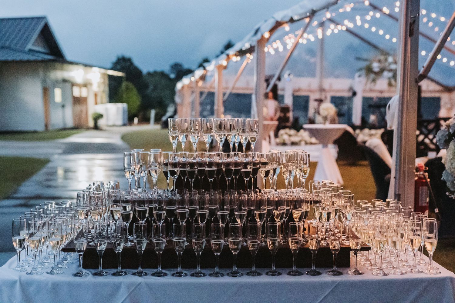 Towering champagne tower with hundreds of glasses arranged in pyramid on table with string lights at dusk.